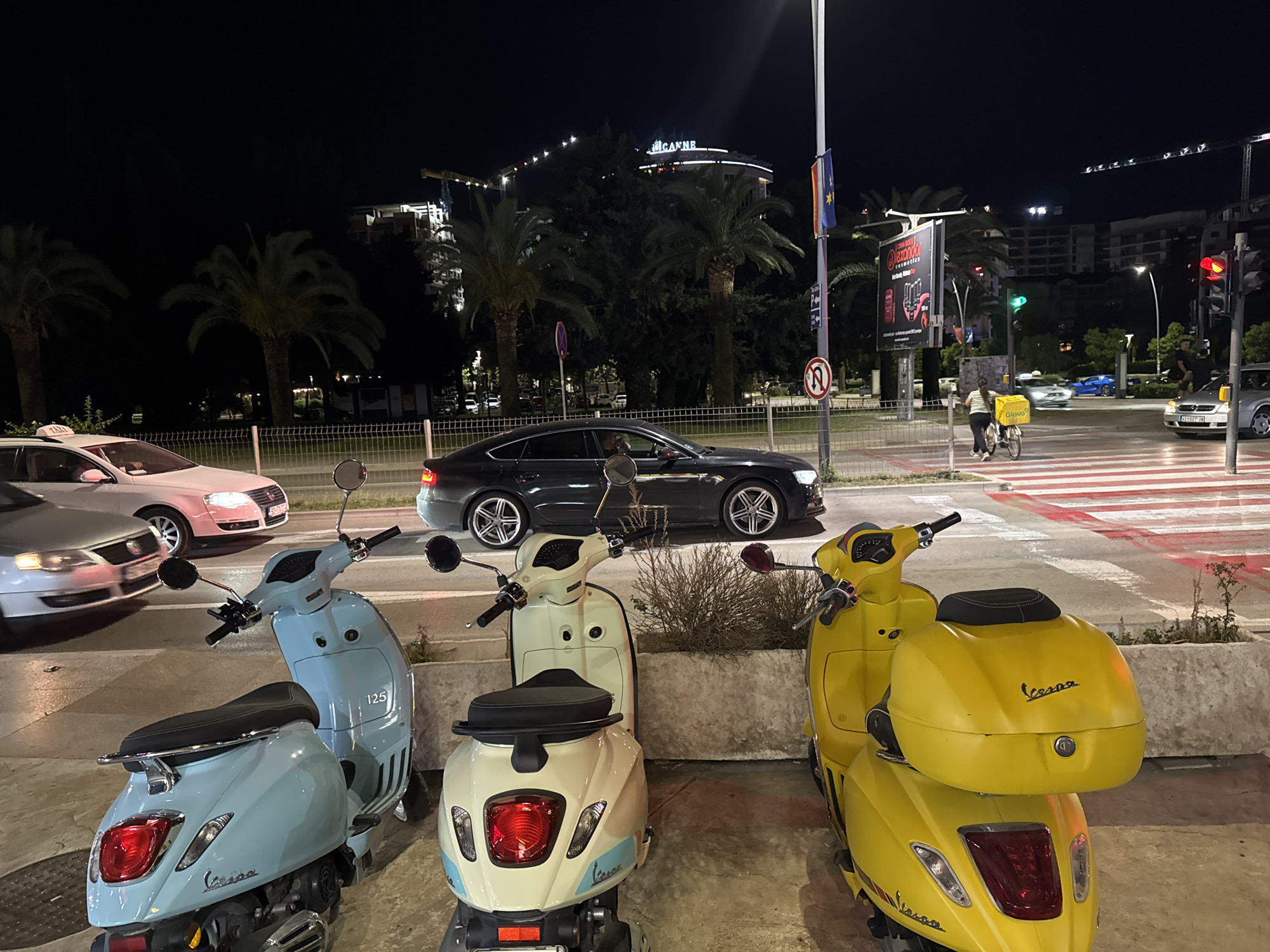 The image shows a street scene at night in Budva, Montenegro. Three Vespa scooters, one light blue, one cream, and one yellow, are parked in the foreground. Behind them is a road with cars, including a silver sedan and a taxi. A person on a bicycle with a "Glovo" delivery box is crossing the crosswalk in the background. There are palm trees and buildings in the distance. A large billboard is on the side of the road. The building in the background has a sign that says "IC ANNE". Traffic lights are visible on the right side of the image.