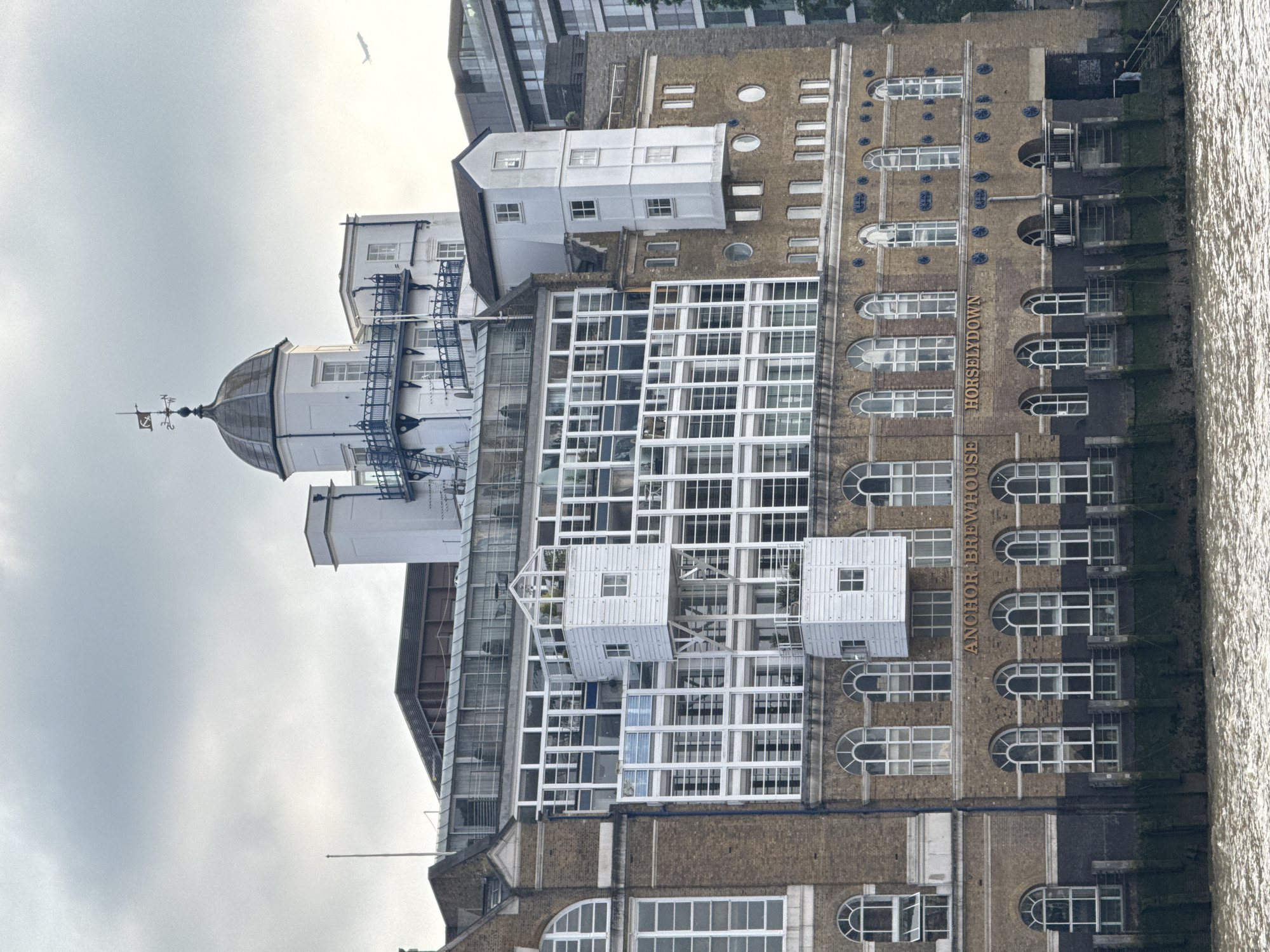 The image depicts the Anchor Brewhouse building in the Horselydown area of London, United Kingdom, situated on the bank of the River Thames. The building exhibits a mixture of architectural styles, including brickwork and modern glass and metal structures. The original brick facade of the building is visible, featuring rows of arched windows and the words "ANCHOR BREWHOUSE HORSELYDOWN" vertically written on its side. Modern additions include a glass-and-metal frame structure and several small, white, house-like extensions affixed to the main building. Atop one section is a structure with a dome and a weathervane. The building faces towards the river, which is visible on the right side of the image. The sky is overcast.