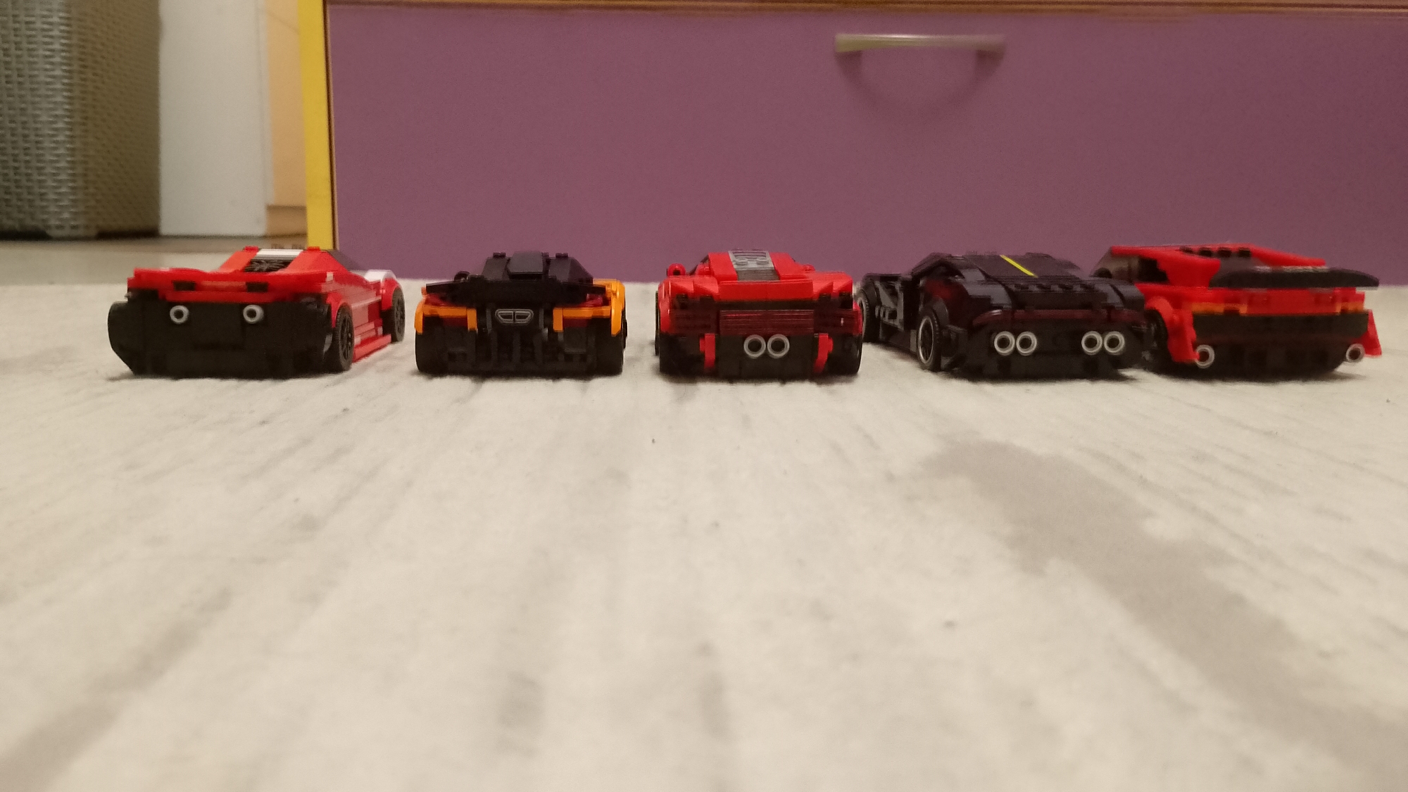 The image displays five brick-built toy cars, likely from a building block set such as Lego Speed Champions, arranged in a horizontal line. All cars are viewed from a low angle, predominantly from their rear. From left to right: the first car is red with black sections and two circular white taillights; the second is black and orange with a rectangular central exhaust; the third, centered, is red and black with two metallic-colored circular exhaust pipes; the fourth is dark gray or black with a distinct yellow stripe and four circular metallic exhaust pipes; and the fifth is red and black, featuring a small rear spoiler and two small circular red taillights.
The cars rest on a light-colored, subtly textured floor, resembling wood or linoleum. The background reveals a domestic interior. On the left, a light-toned wall or furniture piece is partially visible, while on the right, a purple cabinet or drawer unit with a thin yellow horizontal strip and a silver handle is prominent. The cars are stationary, appearing to be posed for display or as if lined up for a race. No text is visible in the image. The scene is an indoor setting, located in Blagoevgrad, Bulgaria.