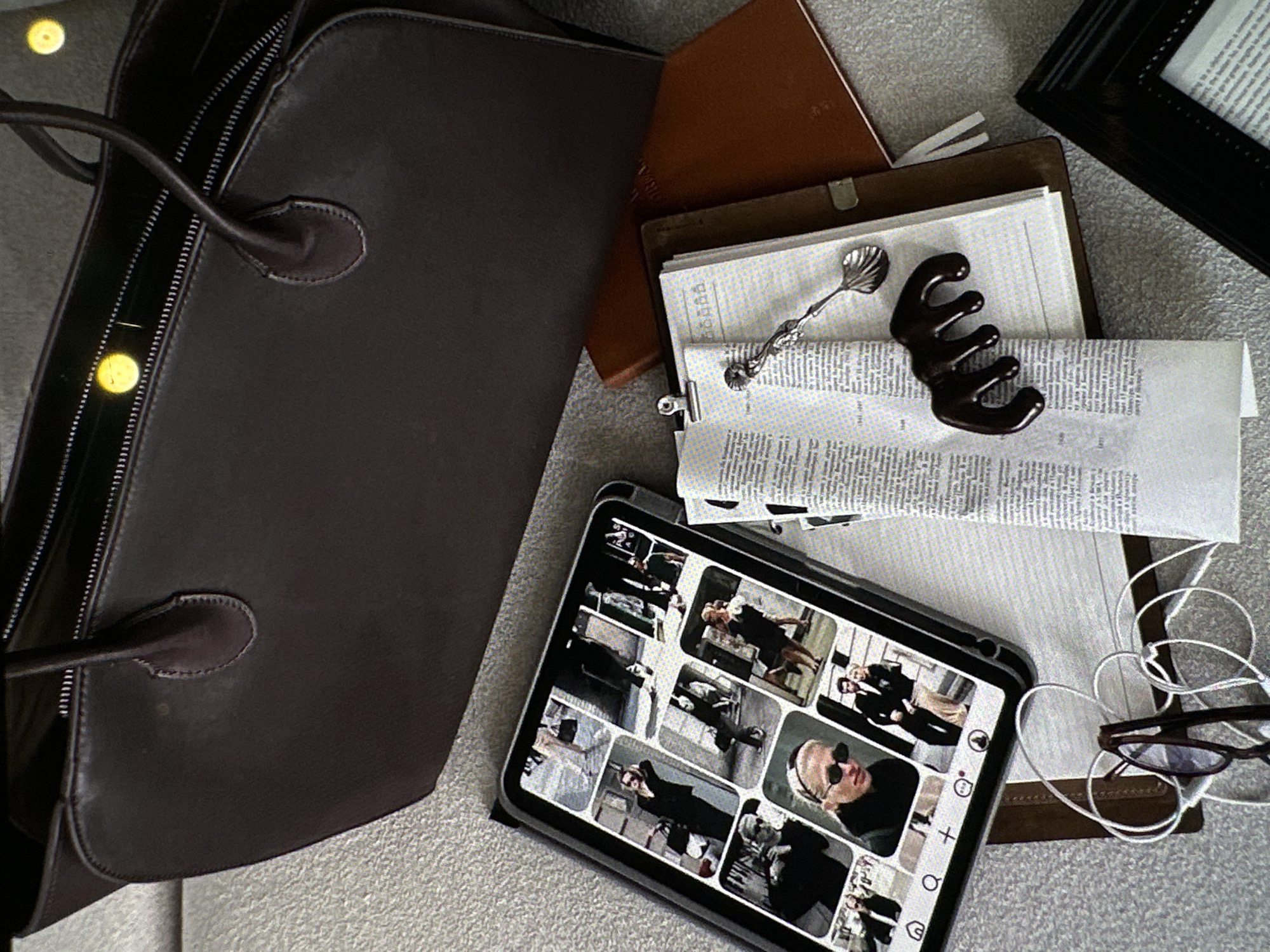 A brown leather bag is positioned on a textured surface, next to a brown notebook, a stack of documents, a silver spoon, a chocolate hair clip, and an electronic tablet. On the tablet screen, black-and-white images are displayed. A pair of eyeglasses with white cords is near the tablet, along with a black frame. The location is not identifiable as Yerevan, Armenia from the visual information provided.