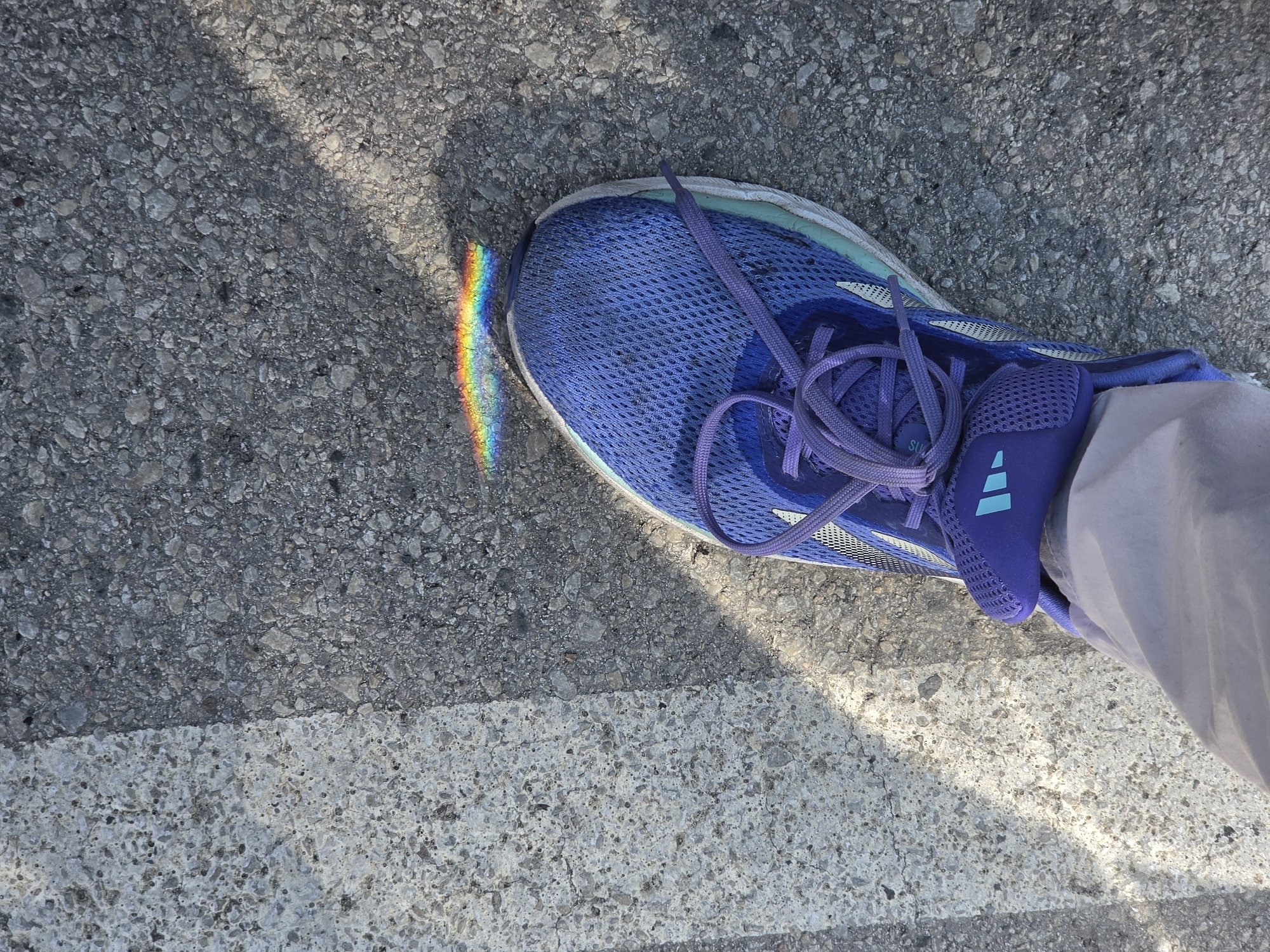 An overhead, eye-level shot depicts a person's left foot wearing a bright blue Adidas running shoe. The shoe is positioned on a textured asphalt surface, which is partially covered by a white painted line that extends vertically on the left side of the frame. A small, iridescent rainbow effect is visible on the asphalt just in front of the shoe, likely caused by light refracting through a prism or water droplet. The shadow of the person's leg and foot is cast diagonally across the asphalt. The overall environment suggests an outdoor urban setting, potentially a street or sidewalk.
