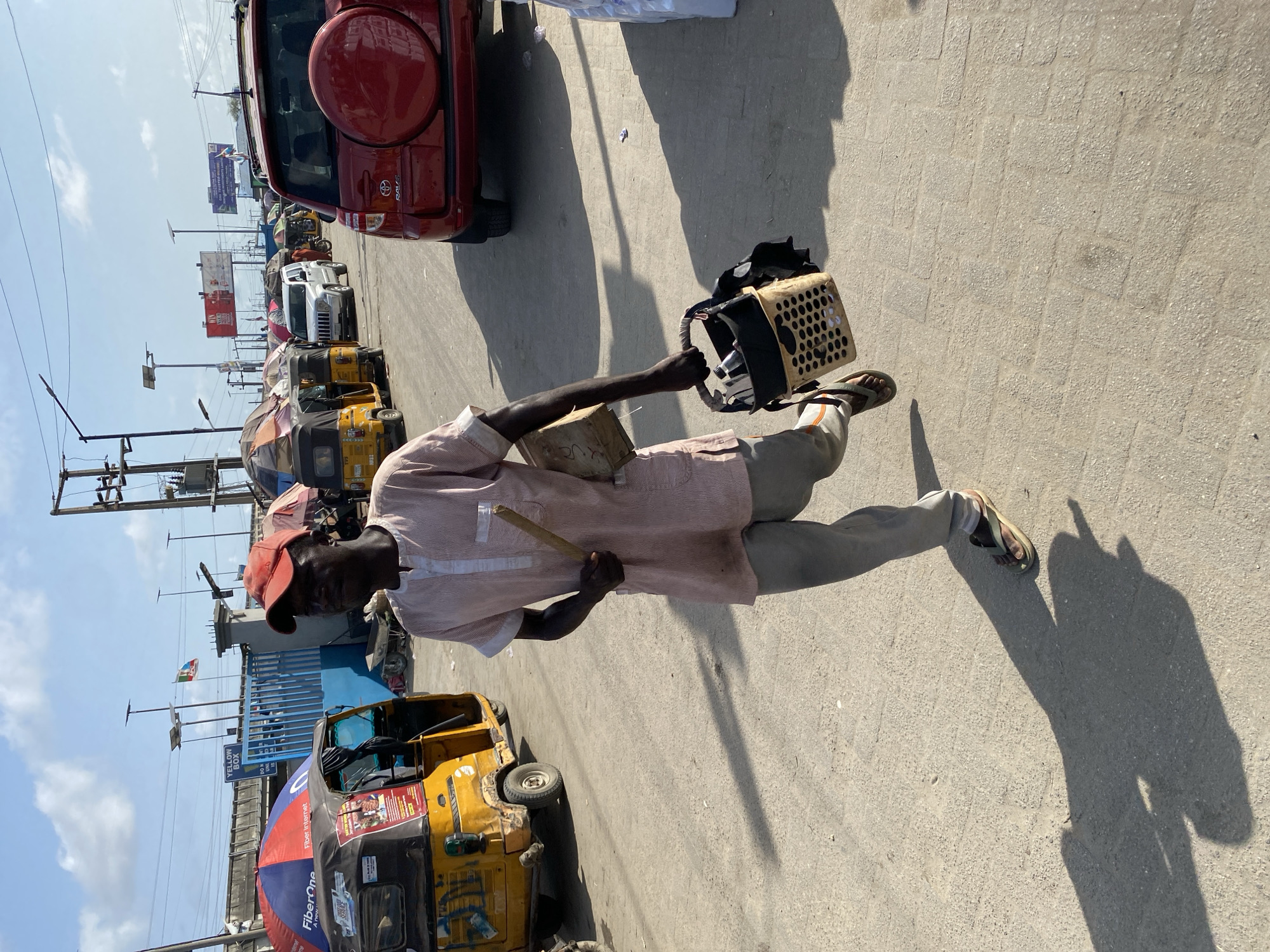 A man walks along a paved street in Lekki, Nigeria, carrying a plastic crate and a wooden object. He wears a red cap, a light pink, striped shirt, and light grey pants, along with sandals. The street is lined with parked vehicles, including red, white, and yellow auto rickshaws. Billboards and utility poles rise in the background. The scene is brightly lit with direct sunlight.