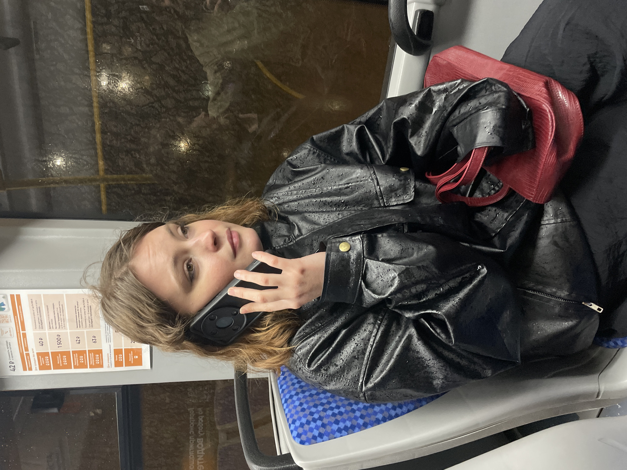 Here's a forensic description of the image:
The image appears to have been taken inside a public transportation vehicle, likely a bus or tram, during or after rainfall. The window shows raindrops. A young woman is sitting, turned towards the camera, and holding a smartphone to her ear. She appears to be engaged in a phone conversation. She is wearing a black, shiny jacket with visible water droplets, suggesting it may be made of leather or a similar water-resistant material. A red handbag is placed next to her on the seat. The seat she is sitting on has a blue patterned fabric.
A sign with ticket prices is visible on the left side of the image. The prices are listed in Russian rubles, which suggests the image was taken in Russia. The name Yekaterinburg is mentioned on the sign. Therefore the image was likely taken in Yekaterinburg, Russia.