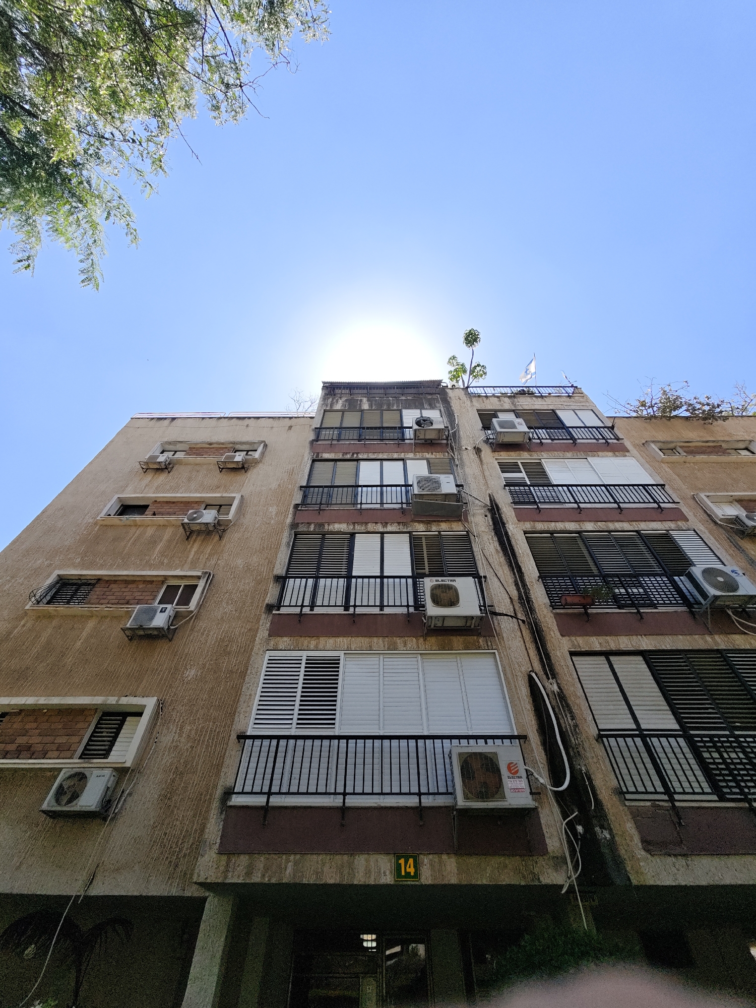 The image shows the exterior of a multi-story apartment building in Petah Tikva, Israel, under a clear blue sky with the sun partially obscured by a tree branch in the upper left. The building's facade is textured and appears weathered, with a mixture of beige and brown paint. Several windows are visible, many equipped with dark metal shutters. Air conditioning units are mounted on the exterior walls. Balconies with black metal railings are present on some floors. A yellow sign with the number "14" is affixed to the lower section of the building. The foreground includes dense green foliage and a glimpse of a dark, covered area at the base of the building. The architectural style suggests an older urban development.