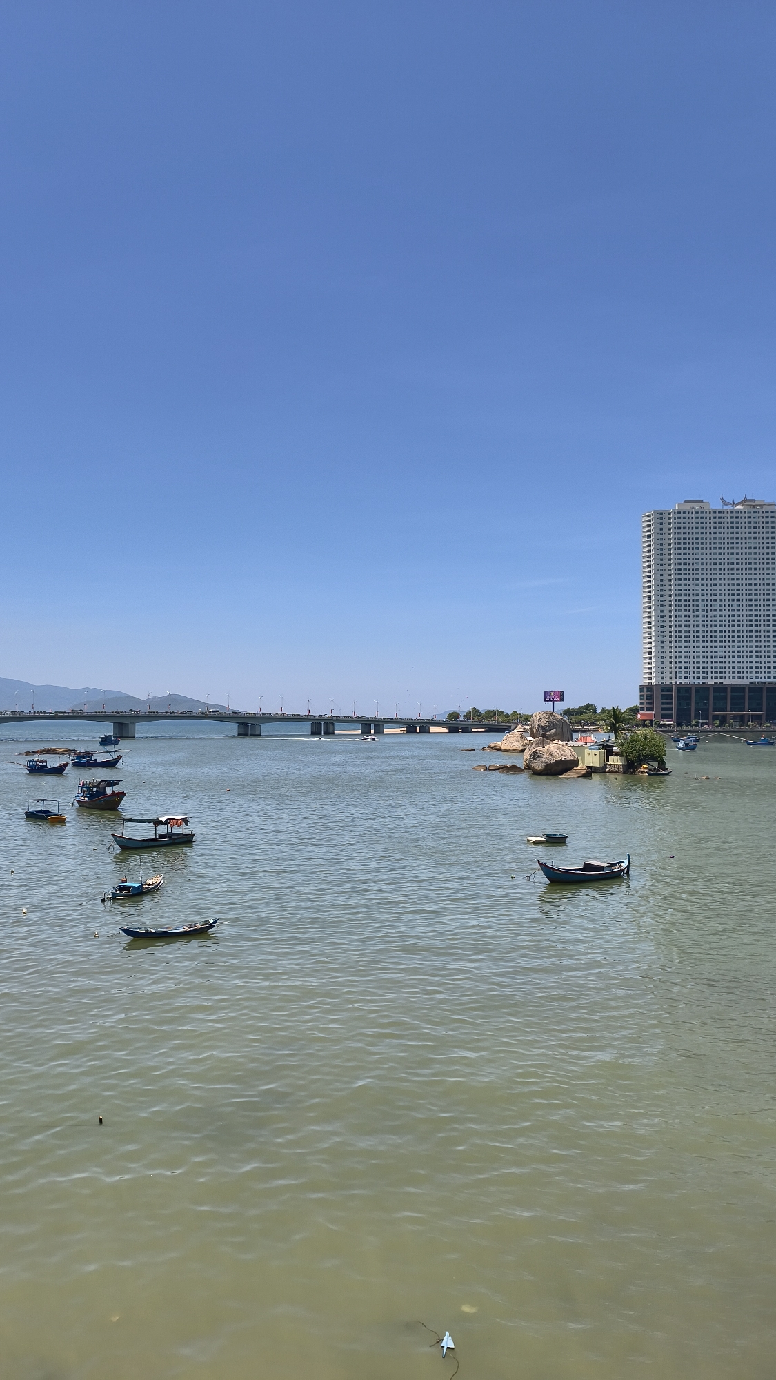 This is an outdoor, eye-level, vertical shot taken on a clear day at the Cai River in Nha Trang, Vietnam. The foreground consists of a body of water with several small boats moored on its surface. In the mid-ground, a long, wide bridge spans the river, with vehicles visible on it, suggesting it's a road bridge. To the right of the bridge, a rocky outcropping with a small structure and a billboard rises from the water. Dominating the right side of the background is a tall, white, modern high-rise building with a patterned facade. The sky is a clear, vibrant blue.