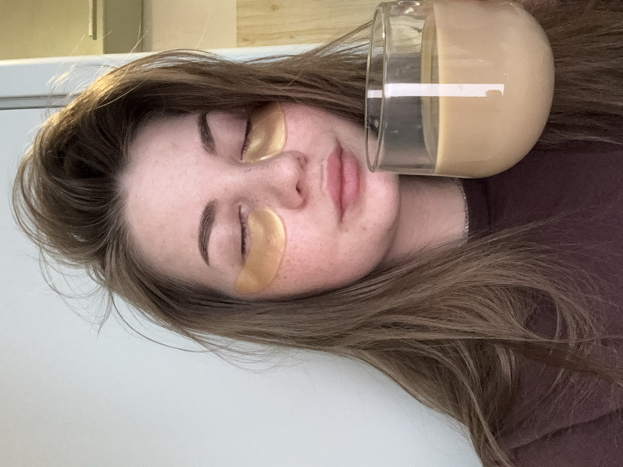 A woman is lying on her side, wearing gold under-eye patches, with a glass of coffee or tea resting near her face. Her eyes are closed and her lips are slightly parted. She has brown hair and freckles. She is wearing a brown shirt. The location is unknown and there is no information in the image or context to determine if this is Chaikovsky, Russia.