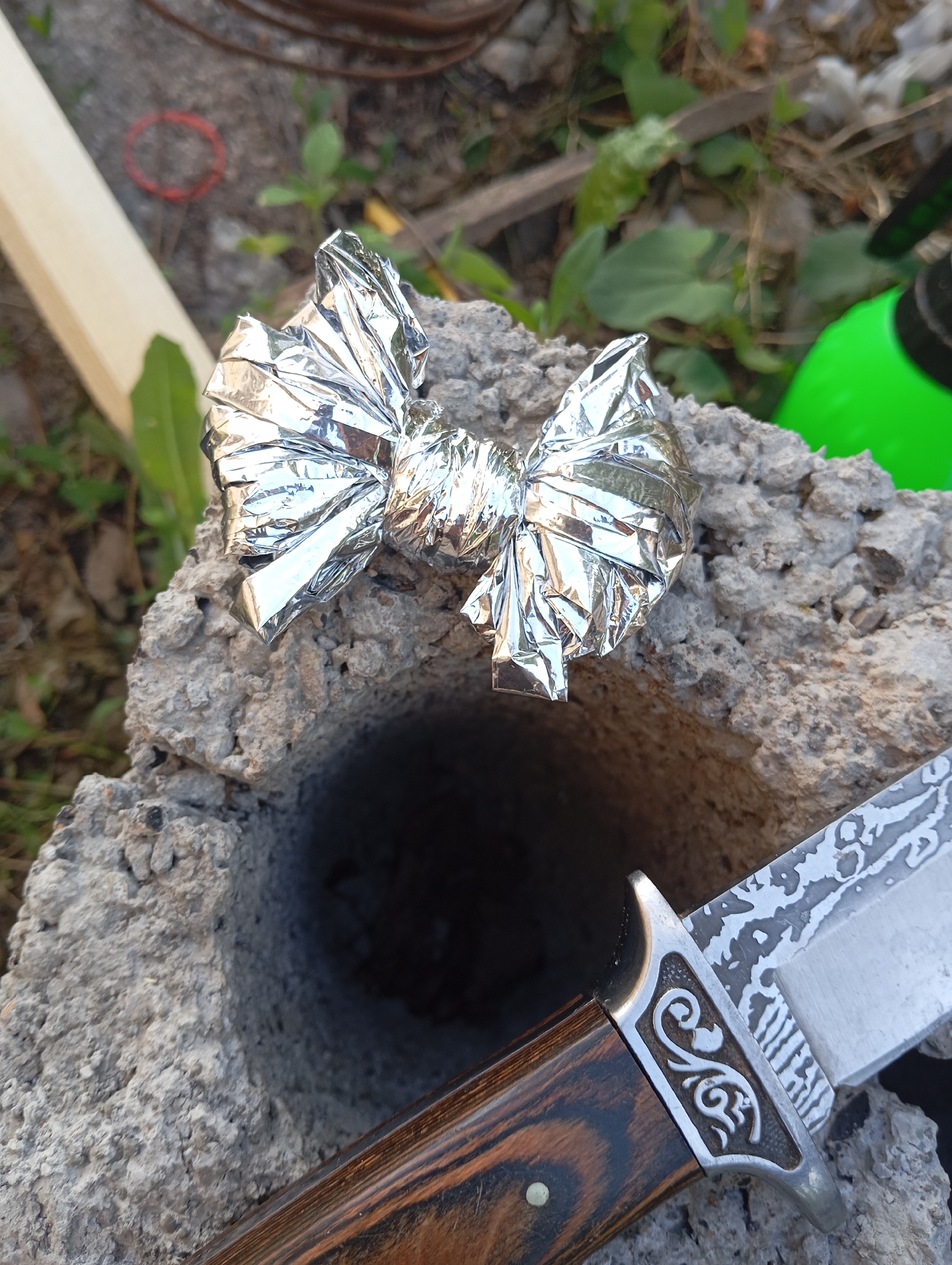This media file shows a close-up, outdoor shot of a bow tie-shaped object made of crumpled aluminum foil, resting on a rough, grey concrete block with a hole in it. To the right of the concrete block, the handle and part of the blade of a knife are visible. The knife has a decorative metal bolster with a swirling pattern and a wooden handle with a visible grain. The background is blurred and shows dry dirt, some green foliage, and what appears to be a coil of dark-colored wire. A bright green object, possibly a spray bottle, is also visible in the upper right background. The lighting suggests it is daytime, likely outdoors with natural light. The overall scene is somewhat informal and suggests an outdoor setting, possibly in a yard or garden. There is no visible text in the image. The location context provided is Orenburg, Russia.