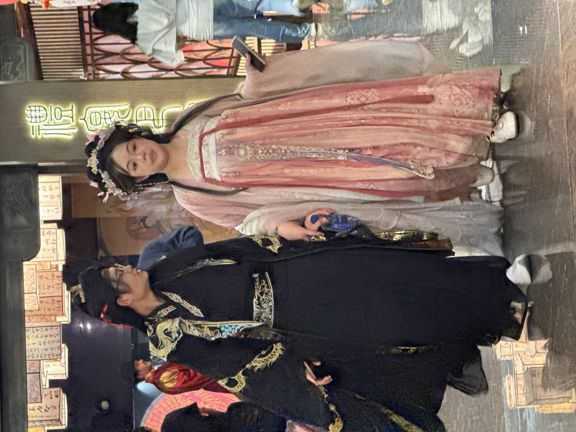 In Jiulongpo District, China, a woman dressed in pink traditional Hanfu clothing is seen standing near a man in black Hanfu clothing. Both individuals are holding mobile phones. The background includes storefront signage, other pedestrians, and decorative lanterns.
