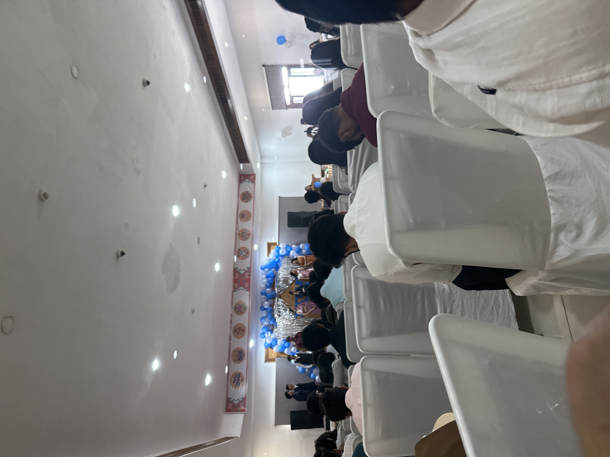 The image shows an indoor event hall, possibly a banquet or conference room. The ceiling is white and has multiple recessed lights and what appear to be security cameras. A banner with circular graphics is displayed above a stage area decorated with blue and white balloons. On the stage, several people are standing, some appearing to be presenting or performing. The foreground is filled with rows of white chairs covered in white fabric, with some attendees seated and facing the stage. The overall atmosphere suggests a formal gathering or celebration.