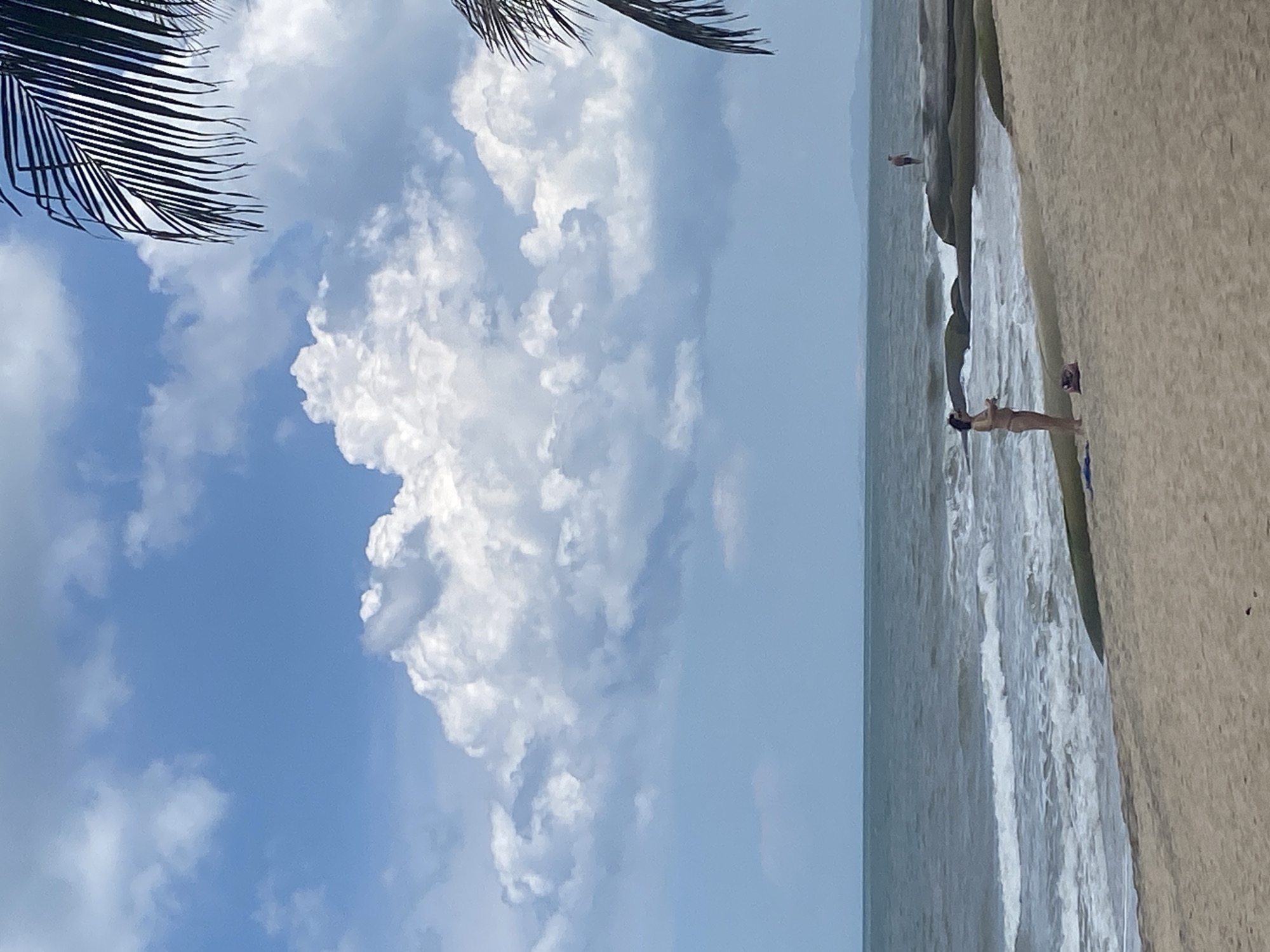 A sunny day at the beach shows a woman in a bikini standing on the sand, facing the water. She is wearing a dark bikini and her hair is dark and short. Her left arm is bent at the elbow and her right arm is down by her side. She has a bag on the sand next to her. In the distance, a man stands on some rocks in the water. Waves crash on the shore and the ocean stretches to the horizon. The sky is blue with large, fluffy white clouds. The sand is light brown and appears dry. A palm frond is visible in the upper right corner of the image, suggesting a tropical location. The scene is one of relaxation and recreation at a coastal area.