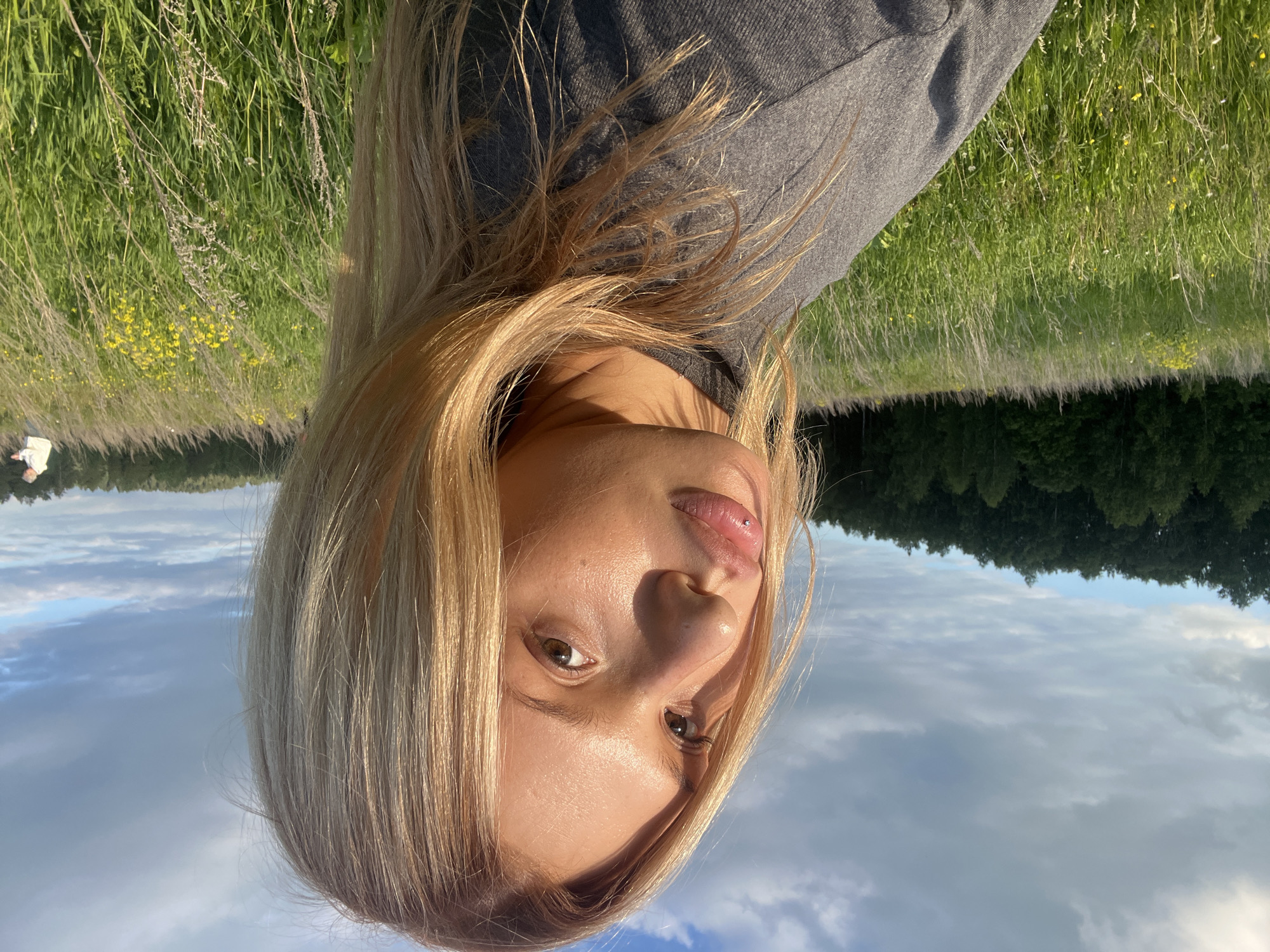 A young woman takes a selfie outdoors in a natural landscape, likely in Bol'shoi Bukor, Russia. The image captures her from a low-angle perspective, with her face prominently featured in the foreground, illuminated by warm, golden light suggestive of late afternoon or early evening. She has long, blonde hair and wears a grey long-sleeved top. Her gaze is directed straight at the camera, and she has a small silver piercing on her upper lip.
Behind her, the background consists of a clear blue sky adorned with white and grey clouds. Below the sky, a distant line of dark green forest creates a distinct horizon. In the immediate foreground, a field of tall green grass with scattered yellow wildflowers is visible, stretching towards the forest line. The lighting highlights her features and hair, creating subtle shadows that add depth to the portrait. The overall scene evokes a calm, natural setting. There is no visible text in the media file.