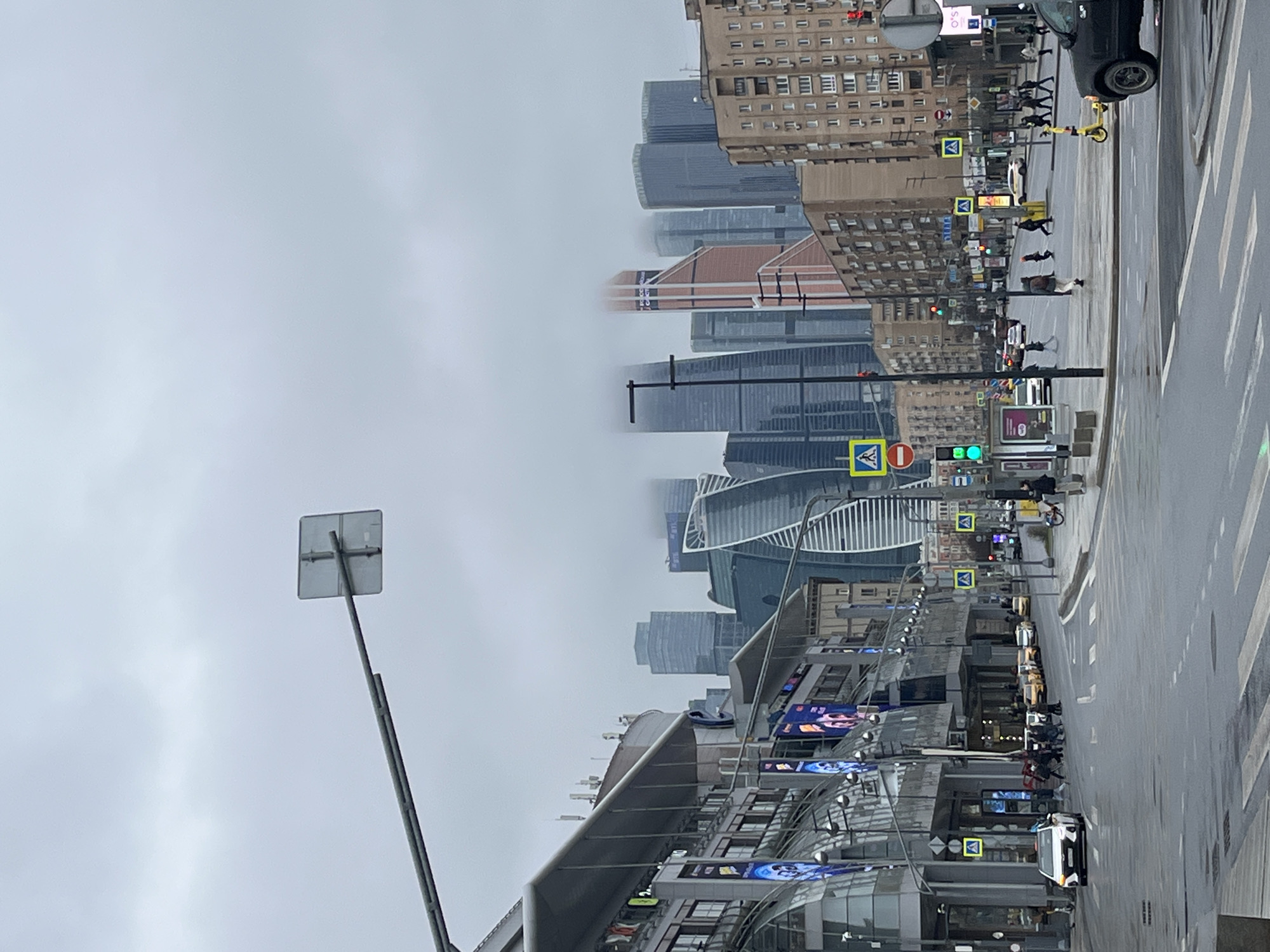 The image depicts a street scene in Moscow, Russia, characterized by dense fog obscuring the upper portions of the city's skyline. Modern skyscrapers and older residential buildings are visible, partially enveloped by the fog. Pedestrians are observed crossing a street with marked crosswalks. Traffic signals and road signs are present. A street lamp or utility pole stands prominently on the left. The street appears wet, possibly due to recent rain or damp conditions.