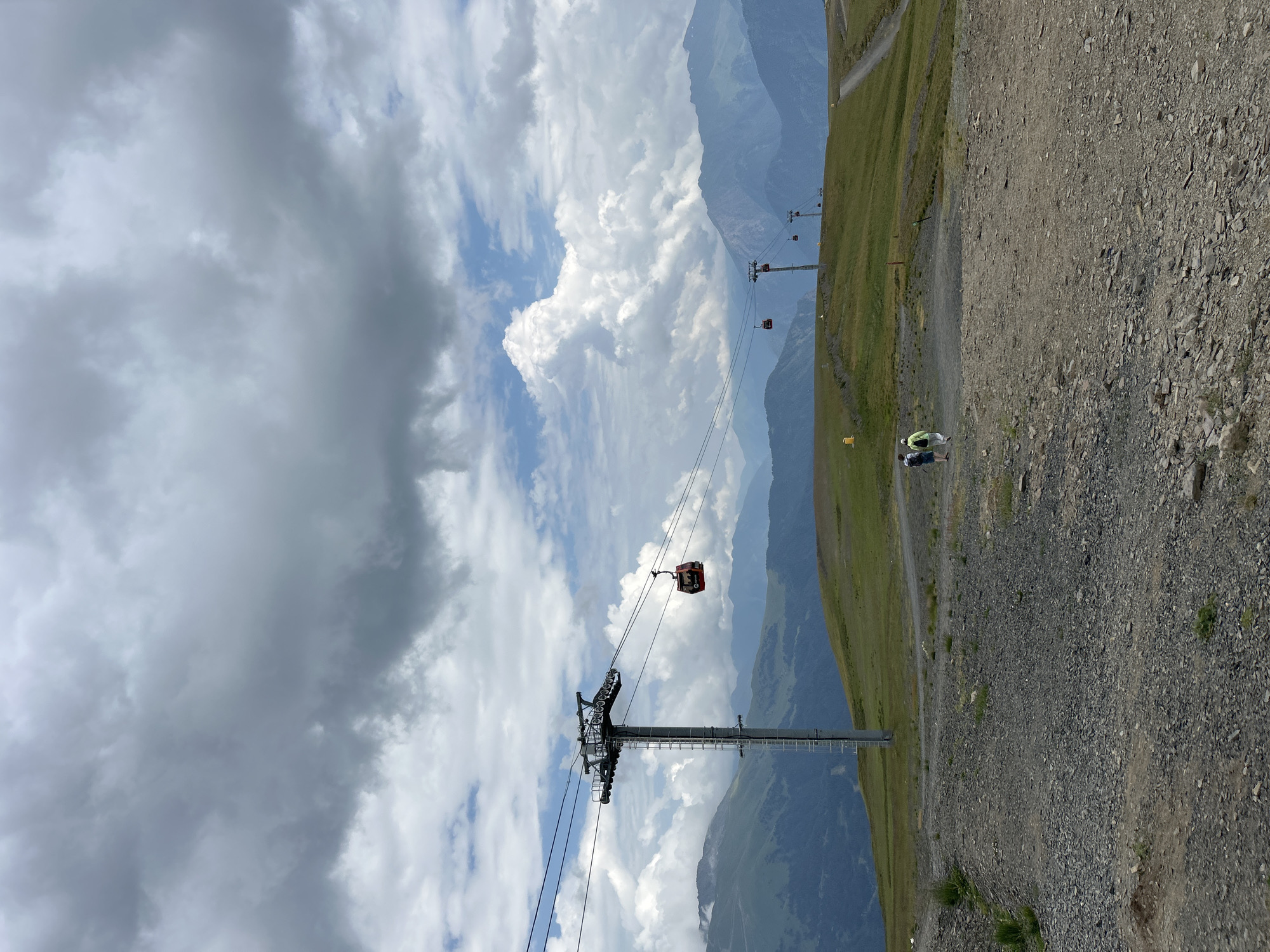 A panoramic view captures a mountainous landscape under a cloudy sky. In the foreground, a gravel path slopes upwards, leading to a grassy incline dotted with two hikers walking away from the camera. A ski lift tower stands prominently on the left, with a red gondola suspended on a cable. Further in the distance, additional ski lift towers and gondolas are visible, traversing the rugged terrain. The background is dominated by layered mountain ranges, shrouded in a hazy blue hue. The sky above is filled with a mixture of dark, heavy clouds and patches of bright blue, suggesting dynamic weather conditions. The scene appears to be set in Lunnaya Polyana, Russia, a location known for its scenic mountainous terrain.