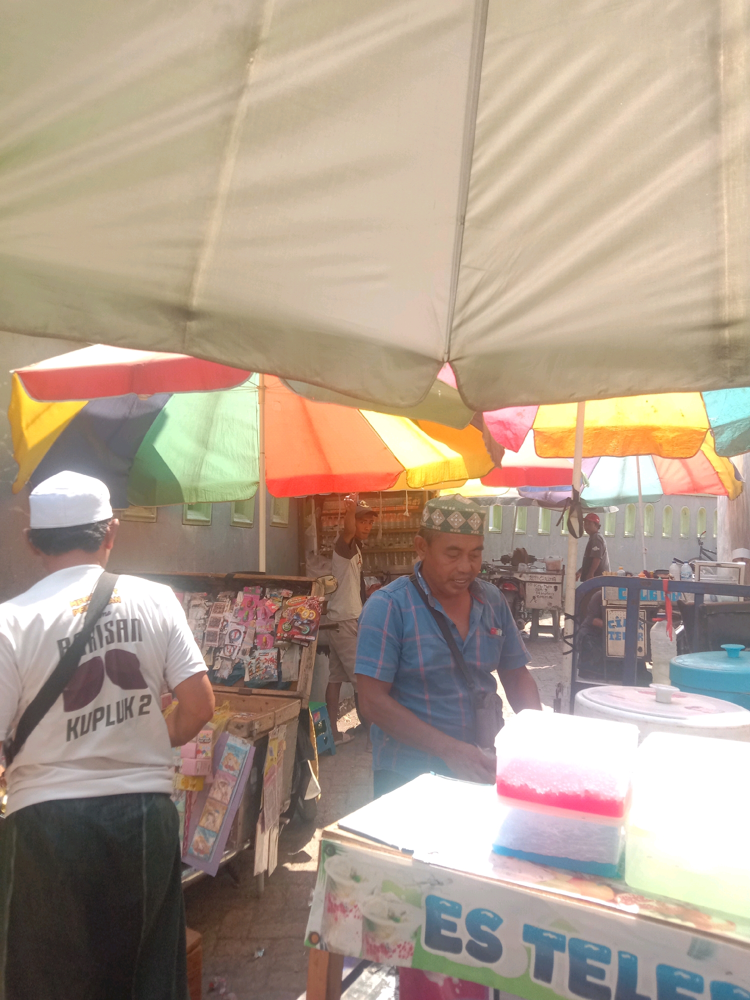 The image depicts an outdoor market scene in Karang Anyar, Indonesia, with several individuals under colorful umbrellas providing shade. In the foreground, a person wearing a white t-shirt with text that includes "KUPLUK 2" and a dark sarong is partially visible from behind, facing away from the camera. To their right, a man in a blue plaid shirt and a patterned kufi cap is looking down, appearing to be engaged in some activity. He has a sling bag across his shoulder. Behind him, another person is visible in a grey shirt and shorts, leaning against a cart filled with various items. Further in the background, more people and market stalls can be seen. The environment is sunny, with a bright light filtering through the umbrellas, casting shadows. There are food or drink containers, possibly for sale, visible on a cart in the right foreground. The overall impression is one of a bustling local marketplace.
