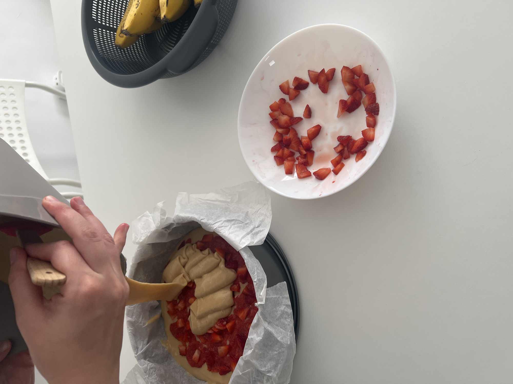 A high-angle shot depicts a cooking scene on a white surface. A black basket containing bananas is located in the upper left. Next to it, there is a white bowl with chopped strawberries. In the lower center, a person is spreading a beige-colored filling on a pie crust, which is covered in chopped strawberries. The pie is in a black baking pan lined with parchment paper. The scene appears to be indoors, possibly in a kitchen. The location is unspecified but presumed to be in the city of Mogán, Spain.