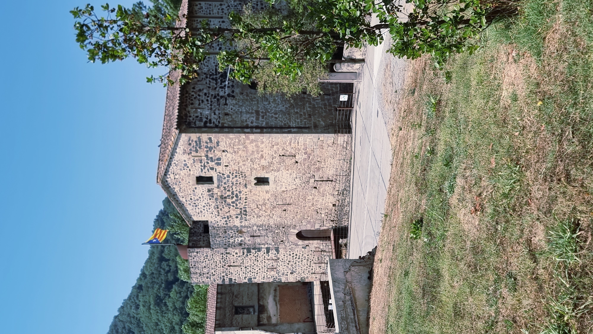 The image depicts the exterior of a stone building, likely a historical structure, situated in Sant Joan Les Fonts, Spain. The building features a tiled roof, small rectangular windows, and a prominent wooden door. A Catalan flag flies from a pole atop a section of the building. To the left of this main structure, another building with a lighter-colored facade and a partially exposed interior is visible. The foreground is dominated by a grassy area that transitions into a paved square in front of the stone building. A tree with green foliage stands to the right, partially obscuring the view of the building. In the background, a lush, tree-covered hillside rises under a clear blue sky. The scene appears to be captured on a sunny day, with the sunlight casting shadows. There are no people visible in the image.