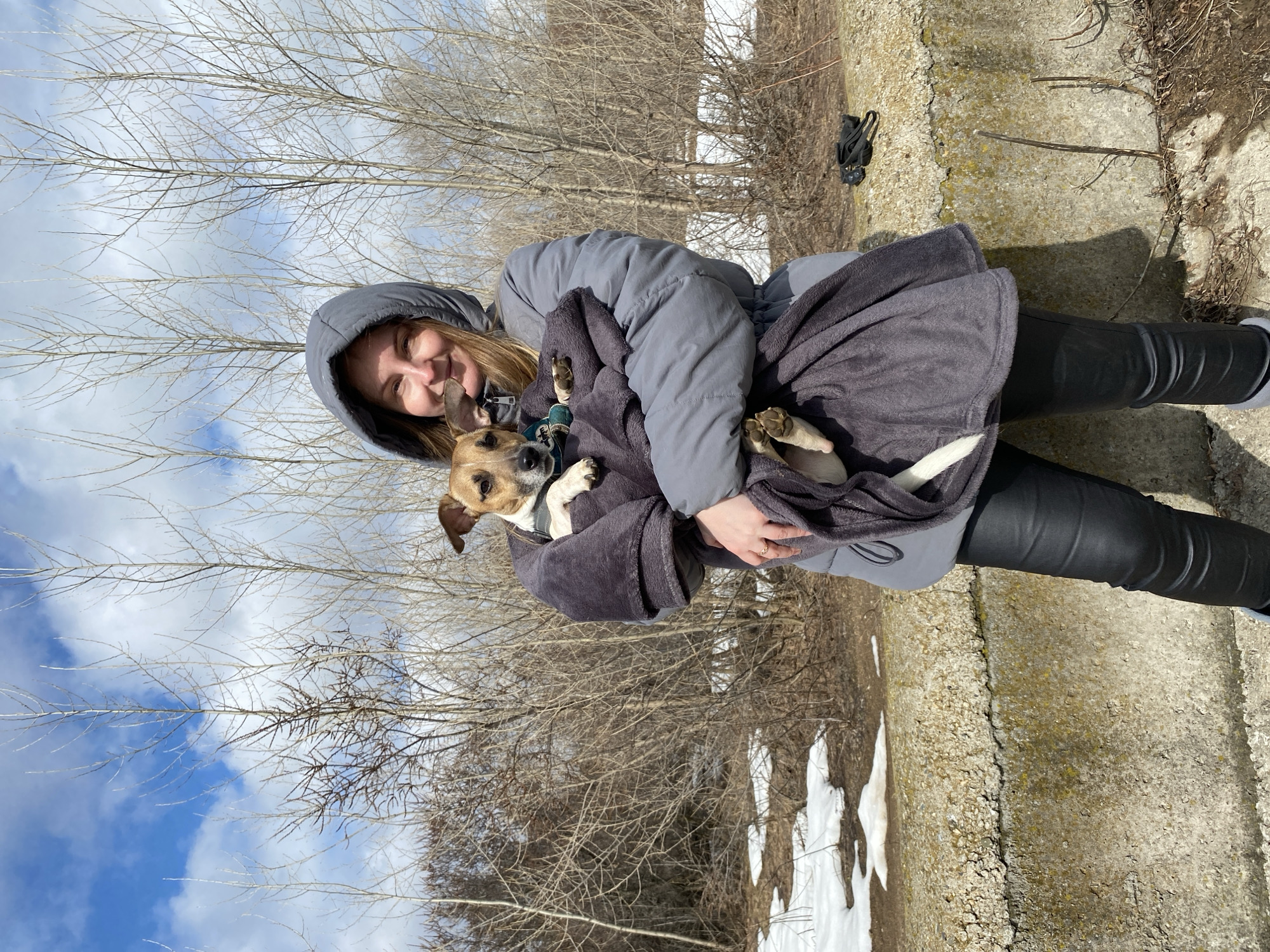 A person wearing a gray hooded jacket and black pants holds a small brown and white dog wrapped in a gray blanket. The person is smiling. The background features bare trees, a blue and white sky, patches of snow, and a concrete structure. Based on the style and location cues, it is likely this image was taken near an urban park or recreational area within the city of Chaikovsky, Russia.