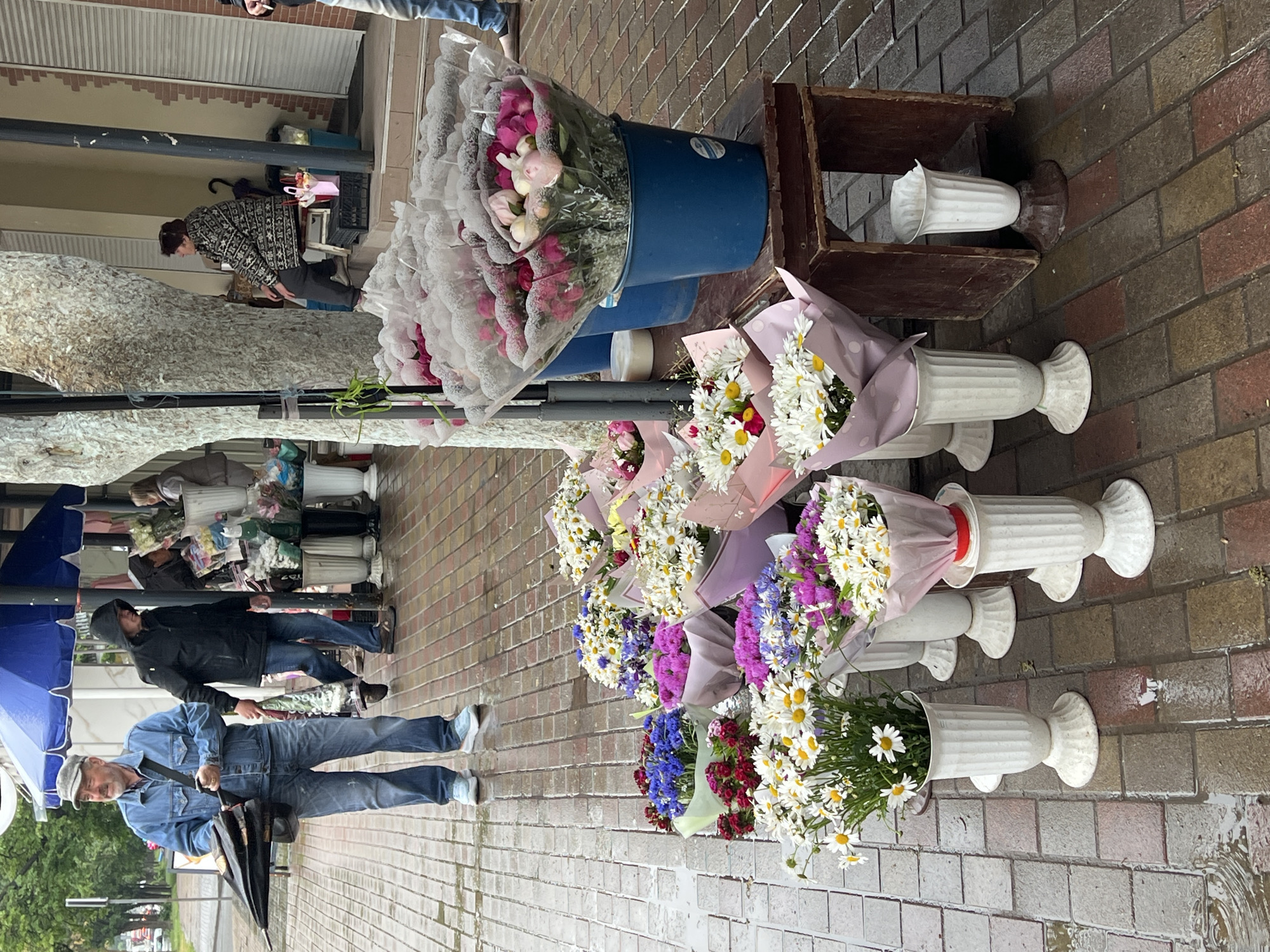This image depicts an outdoor flower market or stall on a wet brick sidewalk in Ternopil, Ukraine. The pavement is visibly damp with puddles, suggesting recent rain.
The main subjects are numerous bouquets of fresh flowers displayed along the sidewalk. These include abundant white daisies, pink and white peonies or tulips, and arrangements of purple, blue, and red blossoms. Many bouquets are wrapped in clear plastic or decorative paper, while some larger bundles are held in two blue plastic buckets. A significant number of bouquets are arranged in tall, white, fluted pedestal vases lining the path.
Several people are present. In the foreground on the left, a mature man with a beard, wearing a blue denim jacket, blue jeans, and a grey cap, stands holding a closed dark umbrella. Next to him, another person in a black hooded jacket and jeans is partially visible. Further back on the right, a woman in a patterned jacket is leaning over a table, seemingly attending to the flower display. More indistinct figures are in the background, suggesting other shoppers or vendors.
The scene takes place during the daytime under what appears to be an overcast sky. The wet ground and the presence of umbrellas indicate cool, possibly damp, weather. The setting is typical of a city street market, with buildings featuring brick walls and roller shutters in the background. No visible text is clearly legible on any signs or objects within the frame. The overall mood is casual and industrious, with no strong emotions expressed by the visible individuals.