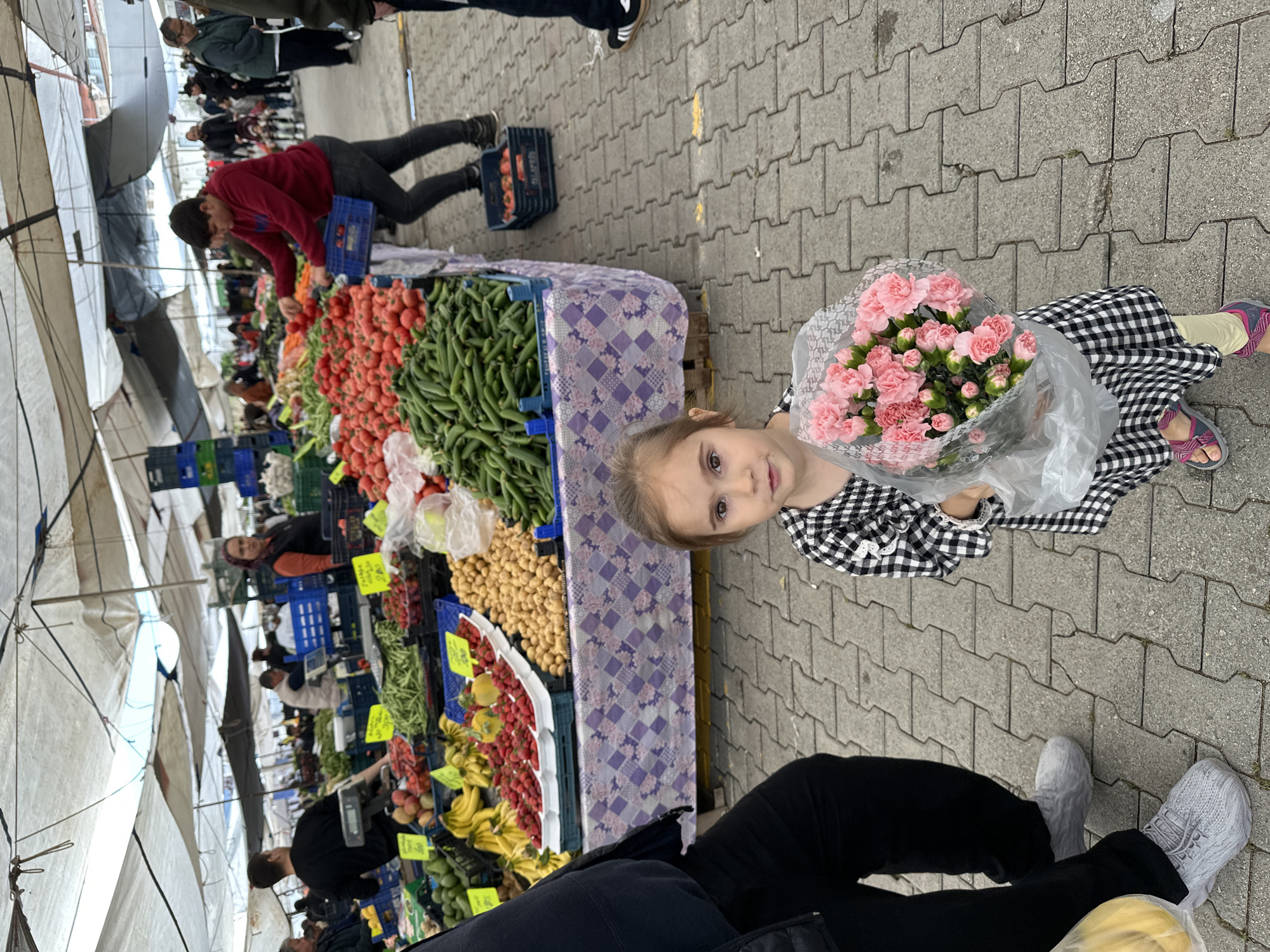 The image shows a young girl holding a bouquet of pink flowers at a market in Fethiye, Turkey. The market is bustling with vendors and customers, with stalls displaying various fruits, vegetables, and other goods. A vendor in a red jacket is picking up tomatoes and placing them into a crate. The ground is paved with interlocking concrete blocks. People are seen walking around the market.