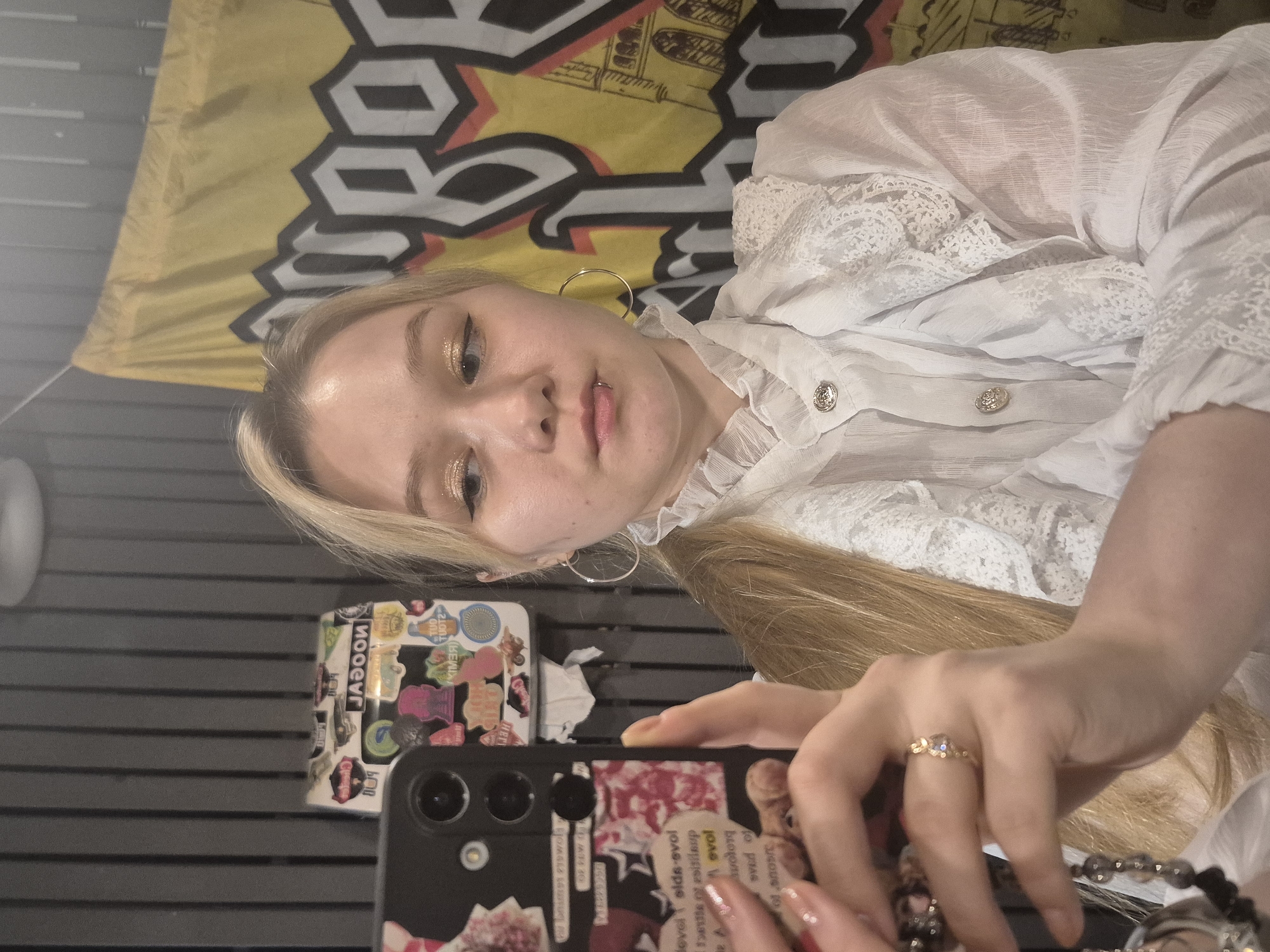 This is a candid, close-up, rotated photo of a young blonde woman. She is looking at the camera, holding her phone in her right hand, which is decorated with stickers and a collage of images. She has glittery eyeshadow and a lip ring. The woman is wearing a white blouse with lace detailing and pearl buttons. A large yellow and black banner with bold lettering is visible behind her. A small stack of items, also covered in stickers, is on a surface to her left. The image appears to be taken indoors, with neutral-toned, textured walls in the background. The lighting is somewhat dim, suggesting it could be evening or an interior space with limited natural light. There is no clear indication of the weather or specific location beyond what is visible in the image. The overall mood is casual and personal, possibly a selfie. Visible text includes "JADOO" and fragmented English words like "levels," "evol," "Fettle," and "estiloup" on stickers.