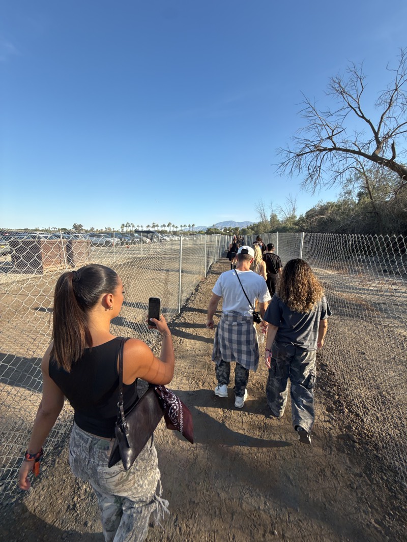 A medium, eye-level shot captures a group of people walking along a fenced path, presumably in Indio, United States. The scene is set outdoors under a clear blue sky, with a few bare tree branches visible on the right.
In the foreground, a woman with a ponytail is holding a phone, appearing to take a photo. She is wearing a black tank top and camouflage pants, with a dark purse slung over her shoulder.
Following her, a group of people walks along the path. A man in a white t-shirt and a plaid shirt tied around his waist is in the lead, followed by another person with curly hair wearing a gray t-shirt and camouflage pants. Other individuals are partially visible further down the path.
On either side of the path, a chain-link fence stretches into the distance. To the left of the fence, a parking lot filled with cars can be seen, with palm trees and mountains visible in the background. To the right of the fence, bushes and trees line the path. The ground is sandy and uneven.