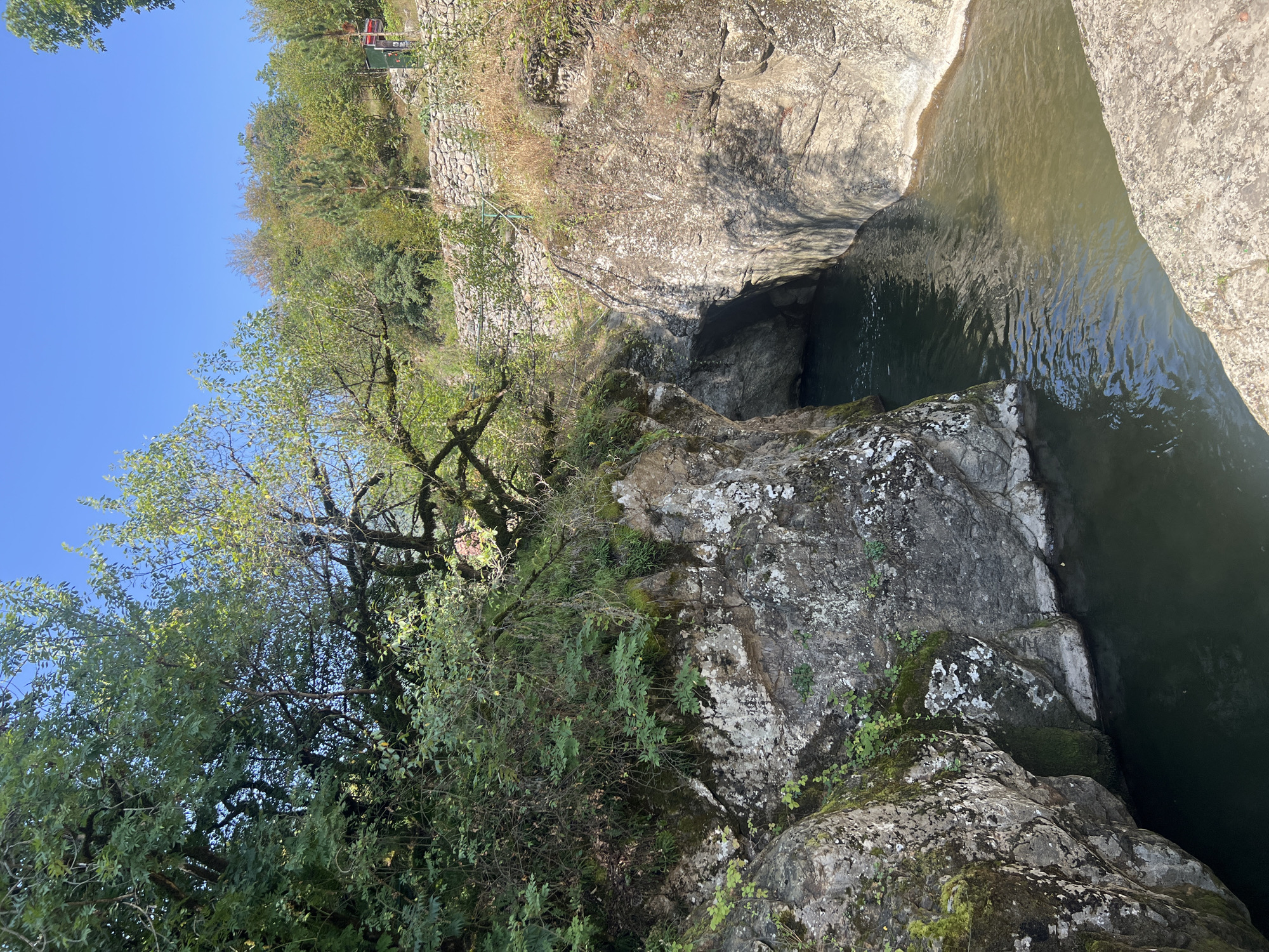 The image depicts a natural gorge with a body of water flowing through it. The gorge is characterized by steep, rocky walls, some of which are covered in moss and vegetation. Trees with green foliage grow along the top edges of the gorge and on the surrounding hillsides. The water in the gorge is dark and appears to be a river or stream. In the background, a dry stone wall runs along a higher elevation, and there is evidence of human presence with a small structure and some greenery visible. The sky is clear and blue. This scene is located in the city of Sarısa, Azerbaijan.