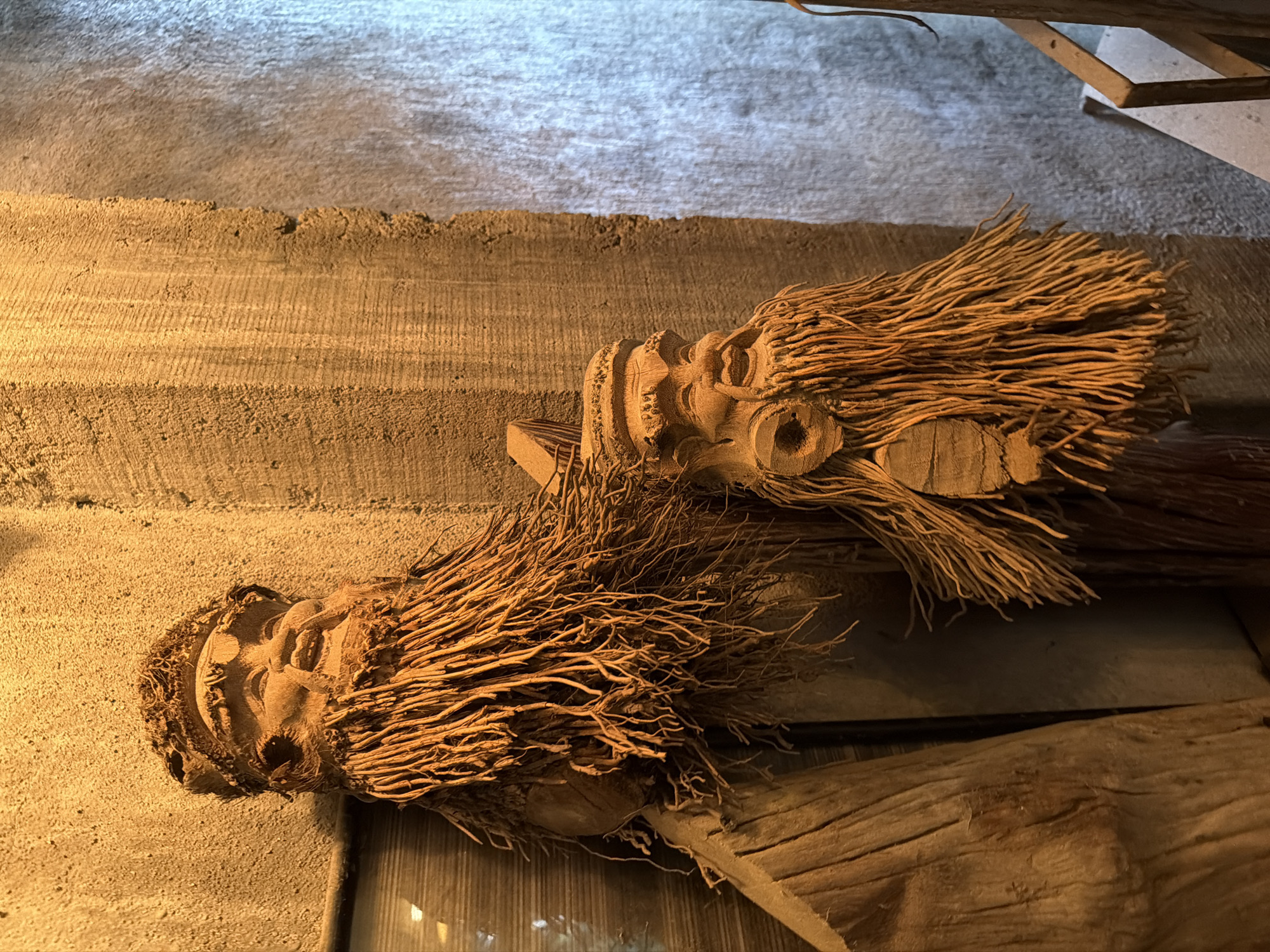 The image shows two carved wooden figures with long, stringy beards, resembling old men with smiling faces. They are positioned in what appears to be an indoor setting with textured walls. The figure on the left is larger and positioned higher than the one on the right. Both figures are crafted from a light-colored wood and have a rustic, handmade appearance. The background consists of a rough, yellowish-brown textured wall on the left and a lighter, possibly concrete or stone wall with vertical striations on the right. The lighting is warm and appears to be artificial, casting shadows and highlighting the textures of the wood and walls. There is no discernible indication of the city of Lâm Viên - Đà Lạt, Vietnam, within the image itself.