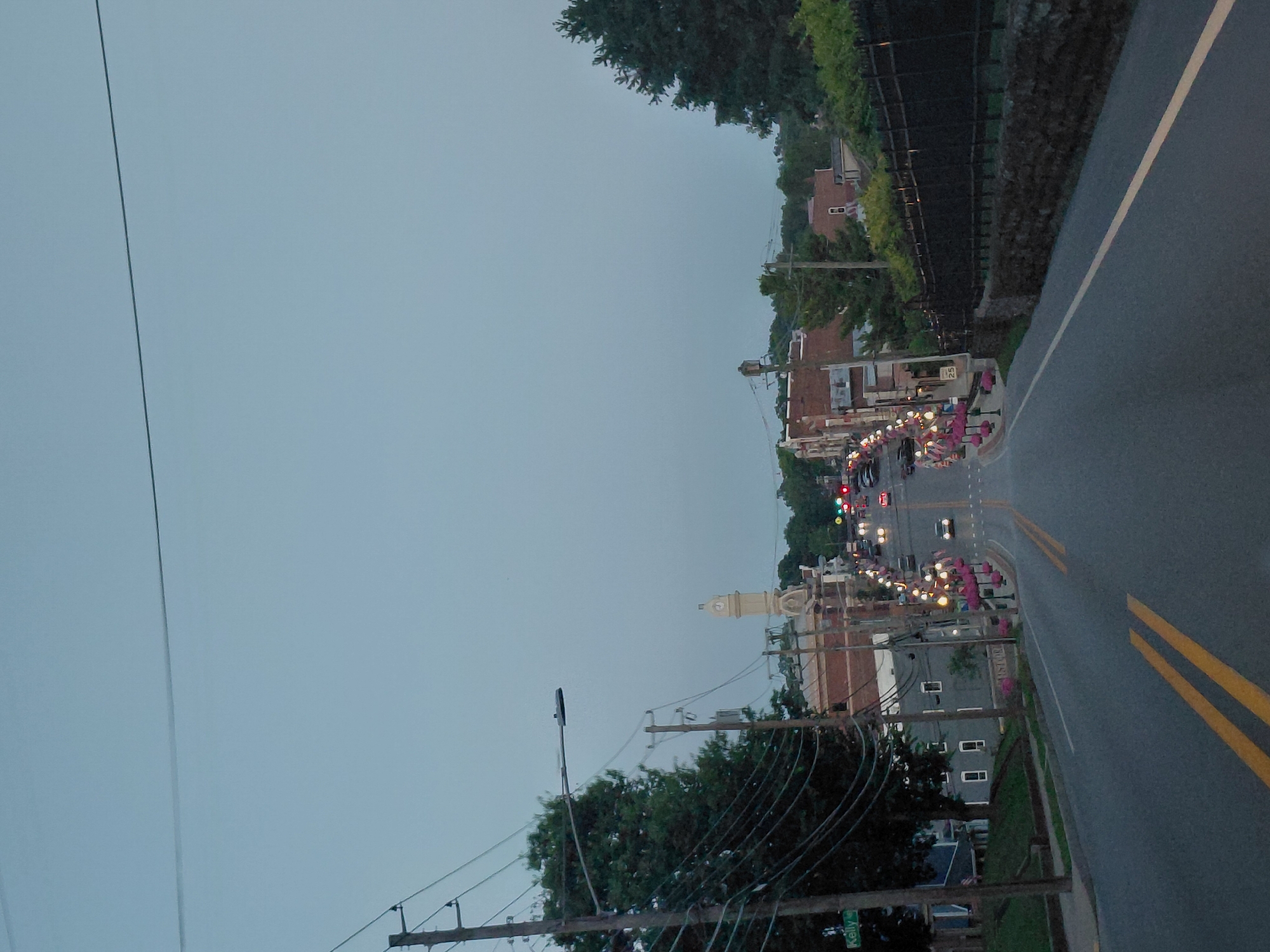 This image captures a street scene in Georgetown, USA, likely during the evening or early morning given the dim, overcast sky and the presence of streetlights. The perspective is from a moving vehicle on a paved road with double yellow lines. The street is lined with buildings on the left, some appearing to be shops or businesses, adorned with festive string lights and planters with pink flowers. A prominent clock tower is visible in the distance. Power lines and utility poles are also a significant feature of the scene. Trees and foliage are present, suggesting a suburban or small-town environment. There are some cars visible on the street, indicating normal traffic. The overall mood is quiet and serene, possibly depicting a town preparing for or winding down from an event. A street sign with "Kelly" is partially visible at the bottom right.