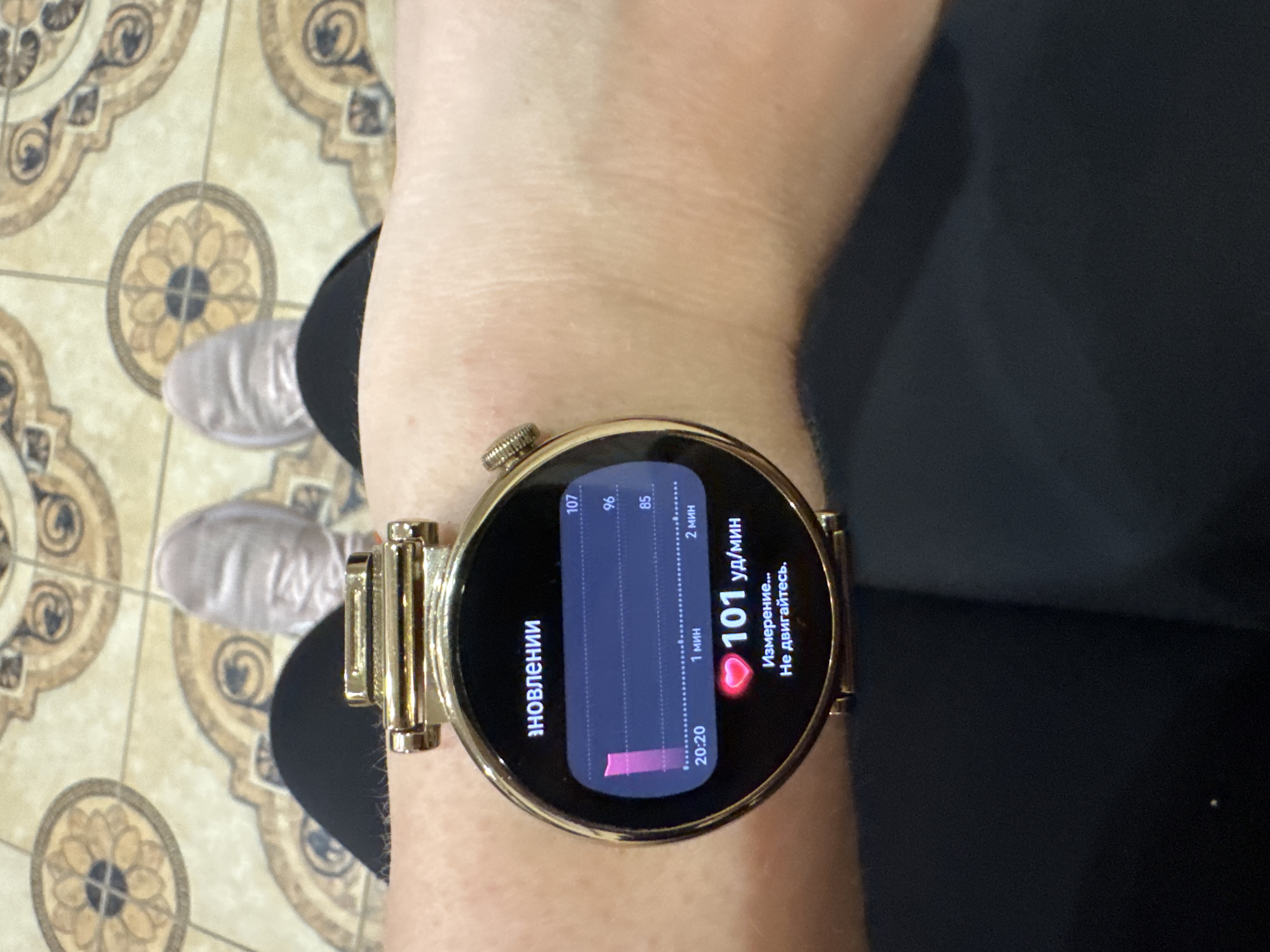 The image features a close-up of a person's wrist wearing a gold-colored smartwatch. The watch display shows a heart rate measurement of 101 beats per minute. The screen also shows a graph depicting heart rate fluctuations over the past two minutes, with readings ranging from 85 to 107. Text in Russian is visible on the screen, indicating it's in the process of measuring and requesting the wearer to stay still. The watch has a metallic gold band, attached to the round face with visible connectors. The person wearing the watch is positioned in front of a patterned floor. The floor is beige with brown circular designs. A portion of the person's clothing is visible, a dark navy or black. The image does not provide information to determine if the location is in Sochi, Russia.