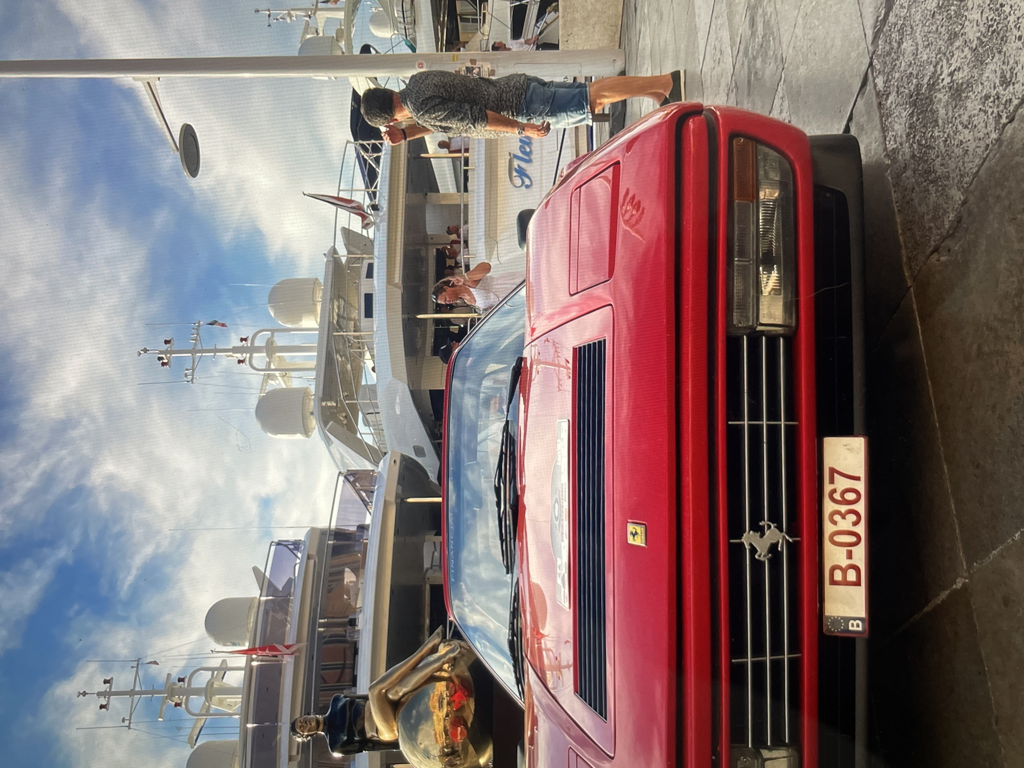Captured in the city of Unknown, Unknown, the image presents a scene where a red Ferrari is parked near a marina filled with yachts. A man stands next to the vehicle, seemingly interacting with the surroundings. Details like the Ferrari's license plate, "B-0367", and the numerous luxury yachts anchored in the background, provide contextual information about the location and potential socioeconomic status. The photograph captures a moment of interaction within an affluent environment.