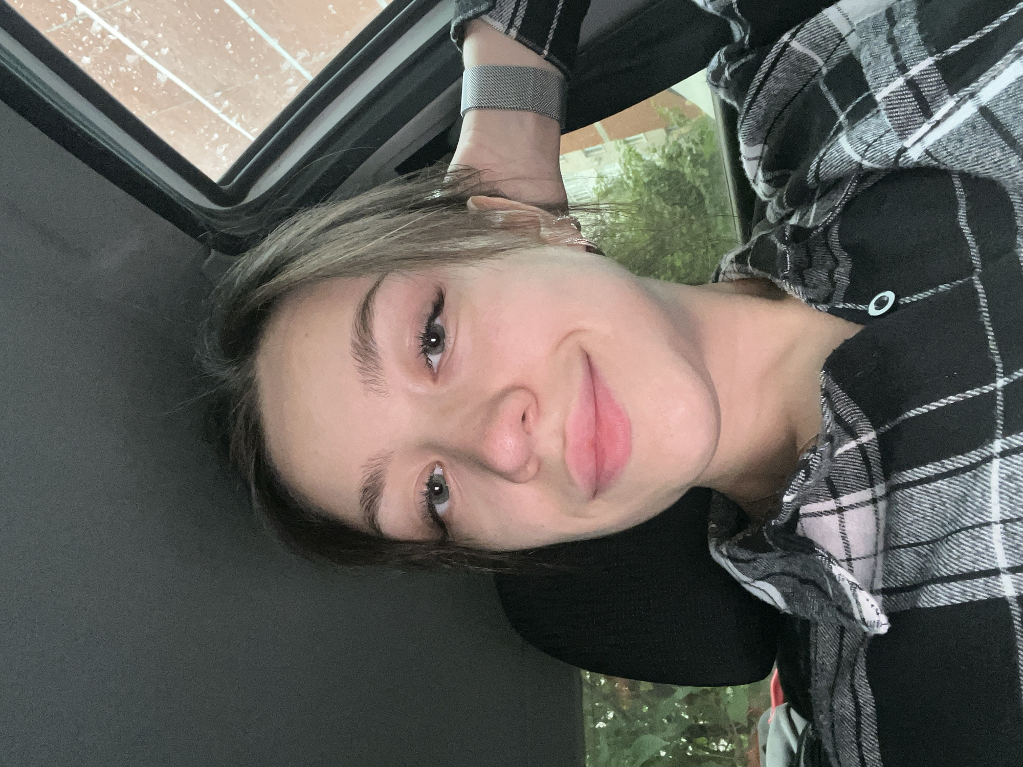 A young woman with light brown hair pulled back is looking at the camera. She is wearing a black and white plaid shirt. Her left arm is bent, with her hand resting on the back of her head. She is inside a vehicle, with the door frame and window visible behind her. Outside the window, a building and trees are visible, suggesting an urban or suburban environment. The scene appears to be in Chaikovsky, Russia, based on the context provided.