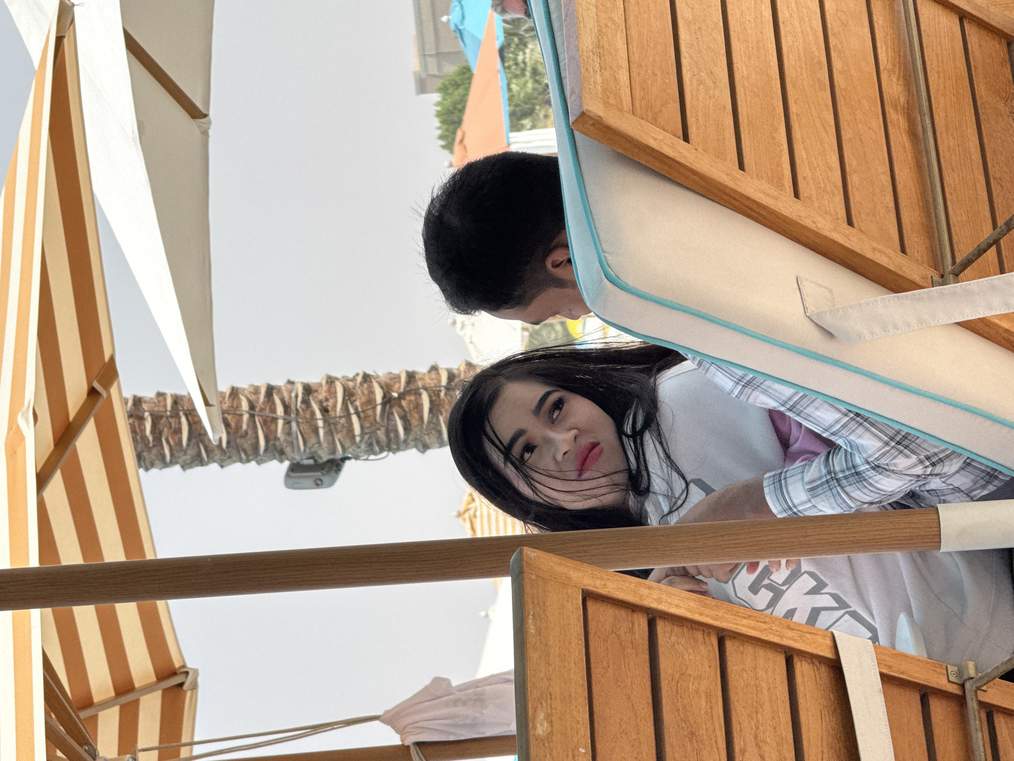 Two individuals are seated on lounge chairs under striped and white umbrellas. The person on the left, a woman with dark hair and red lipstick, is looking to her right. She is wearing a white hoodie with a graphic print and a plaid shirt underneath. The person on the right, a man with dark hair, is facing away from the camera. They are in an outdoor setting, possibly a resort or beach, with palm trees and other umbrellas visible in the background. The scene suggests a relaxing day in a warm climate, given the presence of umbrellas and the bright lighting.