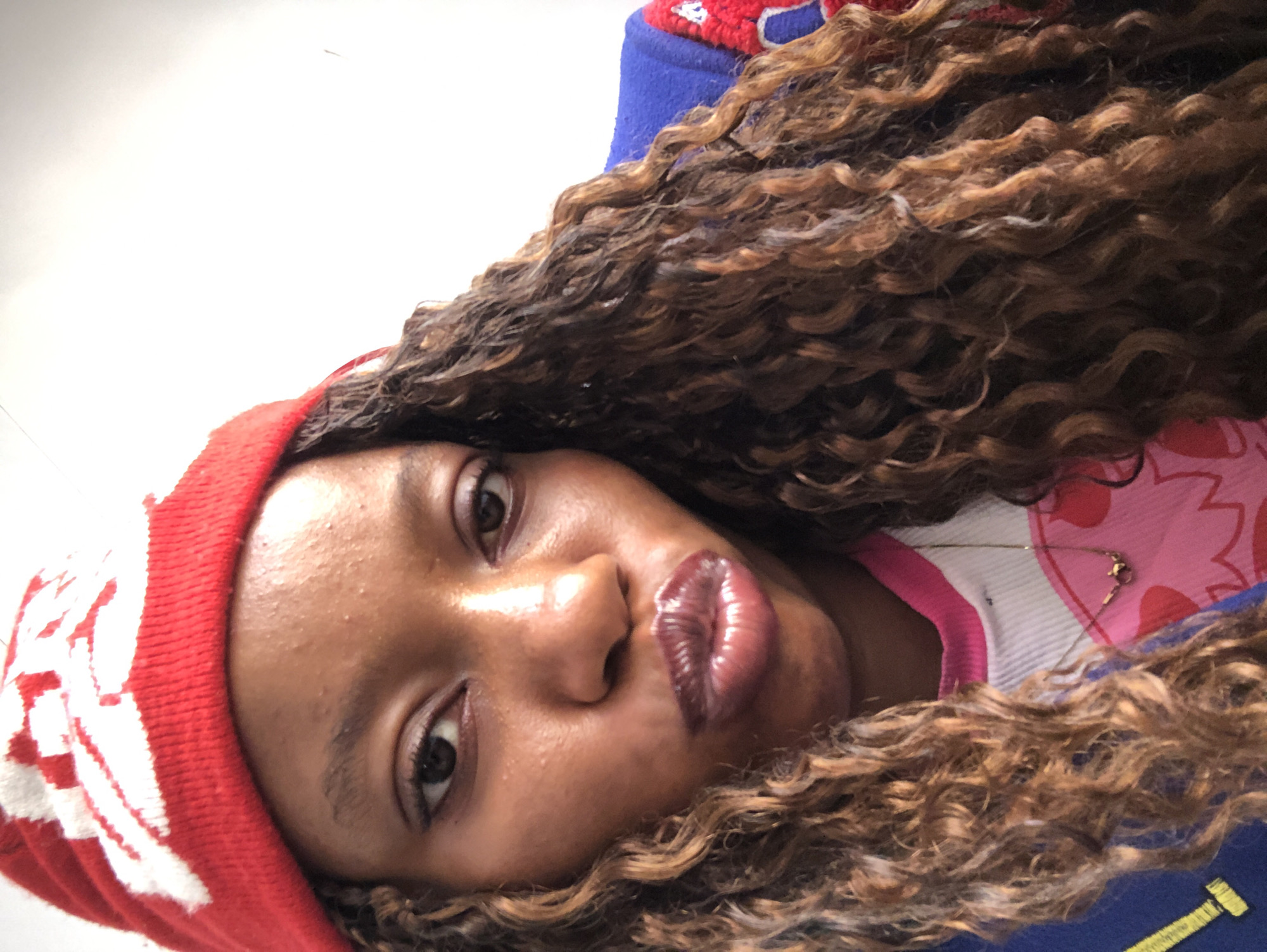A close-up, rotated portrait of a Black woman. She is wearing a red beanie with white designs on the left side of her head, and her brown curly hair frames her face. She is wearing a blue and red jacket over what appears to be a white and pink shirt. A thin gold chain is visible around her neck. The background is white. The location is not identifiable as Pindiga, Nigeria, based on the visual information.