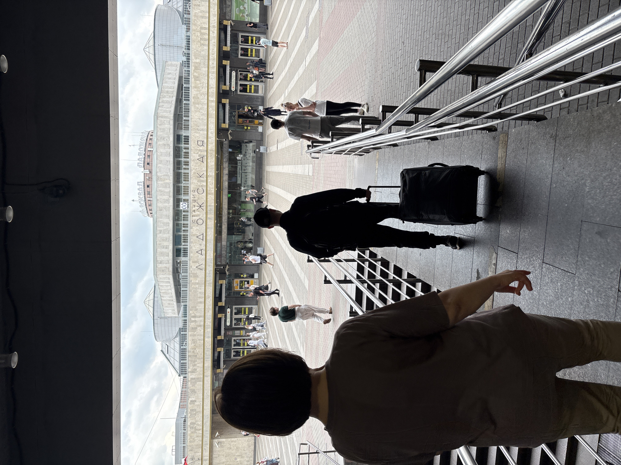 In an outdoor scene at a train station in Moscow, Russia, a woman with short brown hair faces away from the camera, walking down a set of stairs. She wears a brown t-shirt and light-colored pants. In front of her, a man in dark clothing with a baseball cap walks away from the camera, pulling a black wheeled suitcase. Further ahead, several people are visible on a wide, paved concourse in front of a modern building with large glass windows and architectural elements that read "ЛОЖСКAЯ" in Cyrillic. The sky is overcast. The scene appears to be at the Rizhsky railway station.