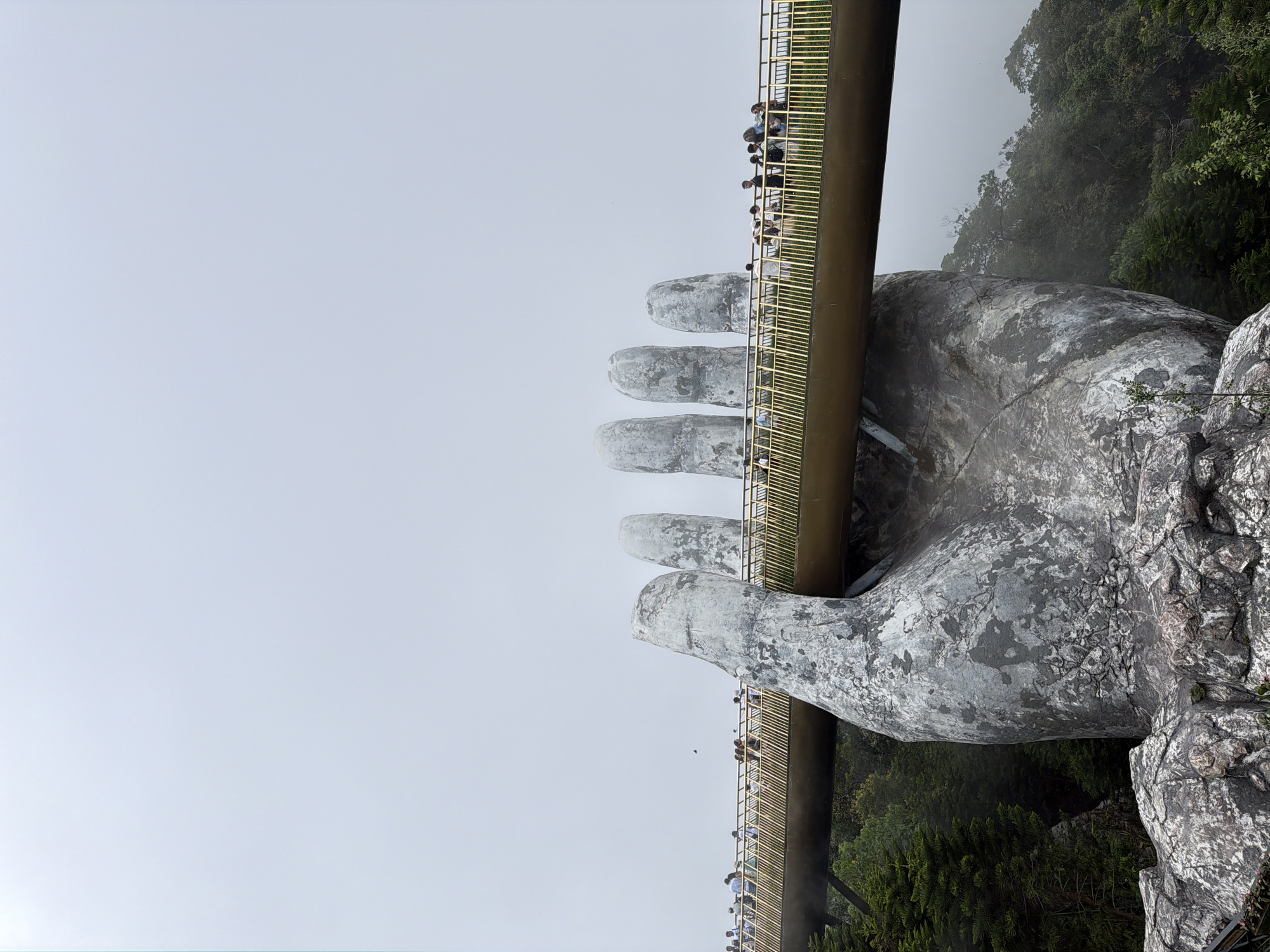 The image shows the Golden Bridge, a pedestrian walkway held up by two giant stone-colored hands, located in the Bà Nà Hills resort near Đà Nẵng, Vietnam. The bridge is filled with people looking at the surrounding landscape. The area is somewhat foggy with a mix of trees and rock formations.