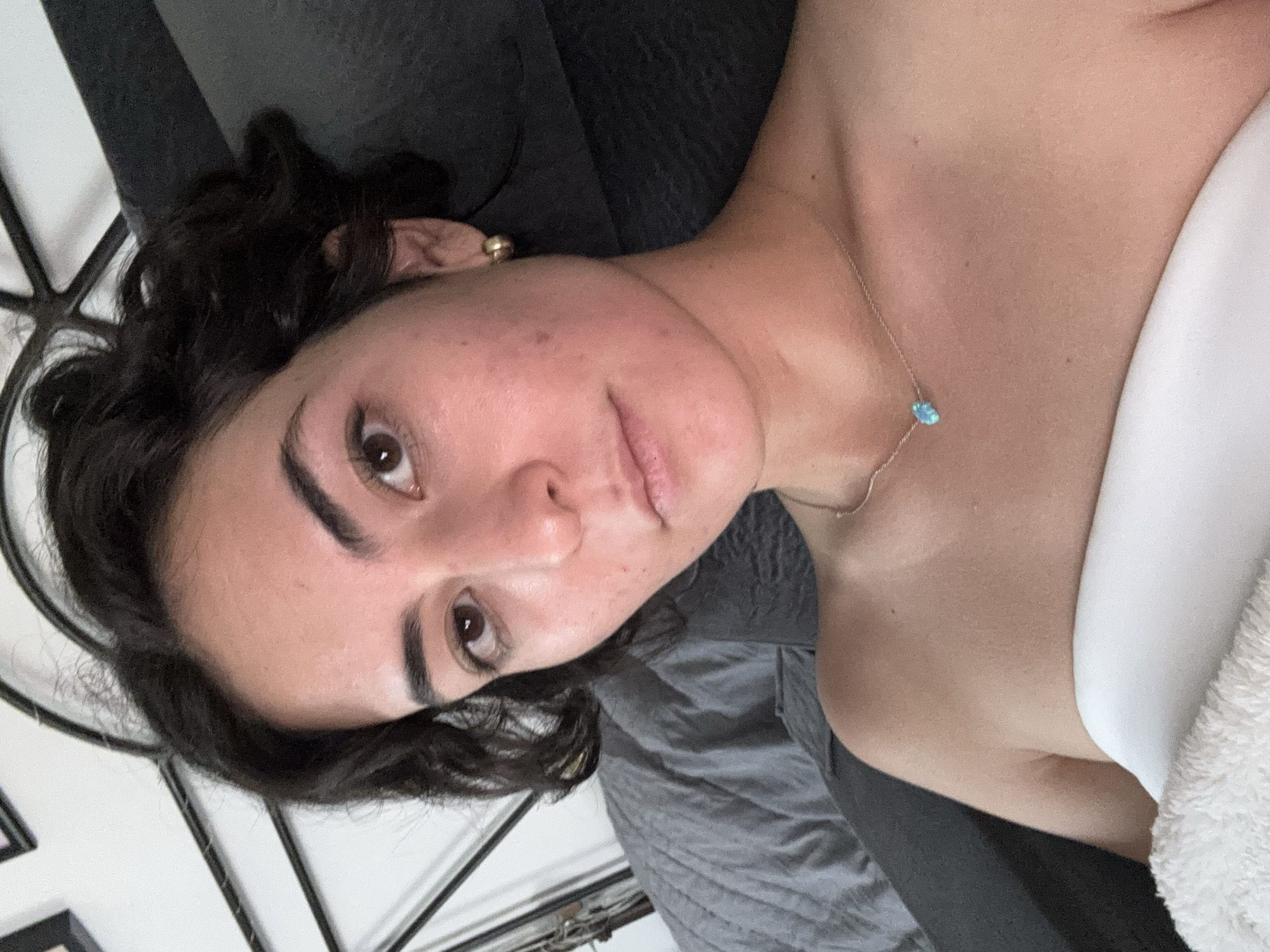 A person with dark curly hair and brown eyes is lying on a bed with a gray blanket and a white towel. They are wearing a silver necklace with a turquoise stone. The person is indoors, as indicated by the bed frame in the background. There is no evidence to suggest the location is in the city of Puebla, Mexico.
