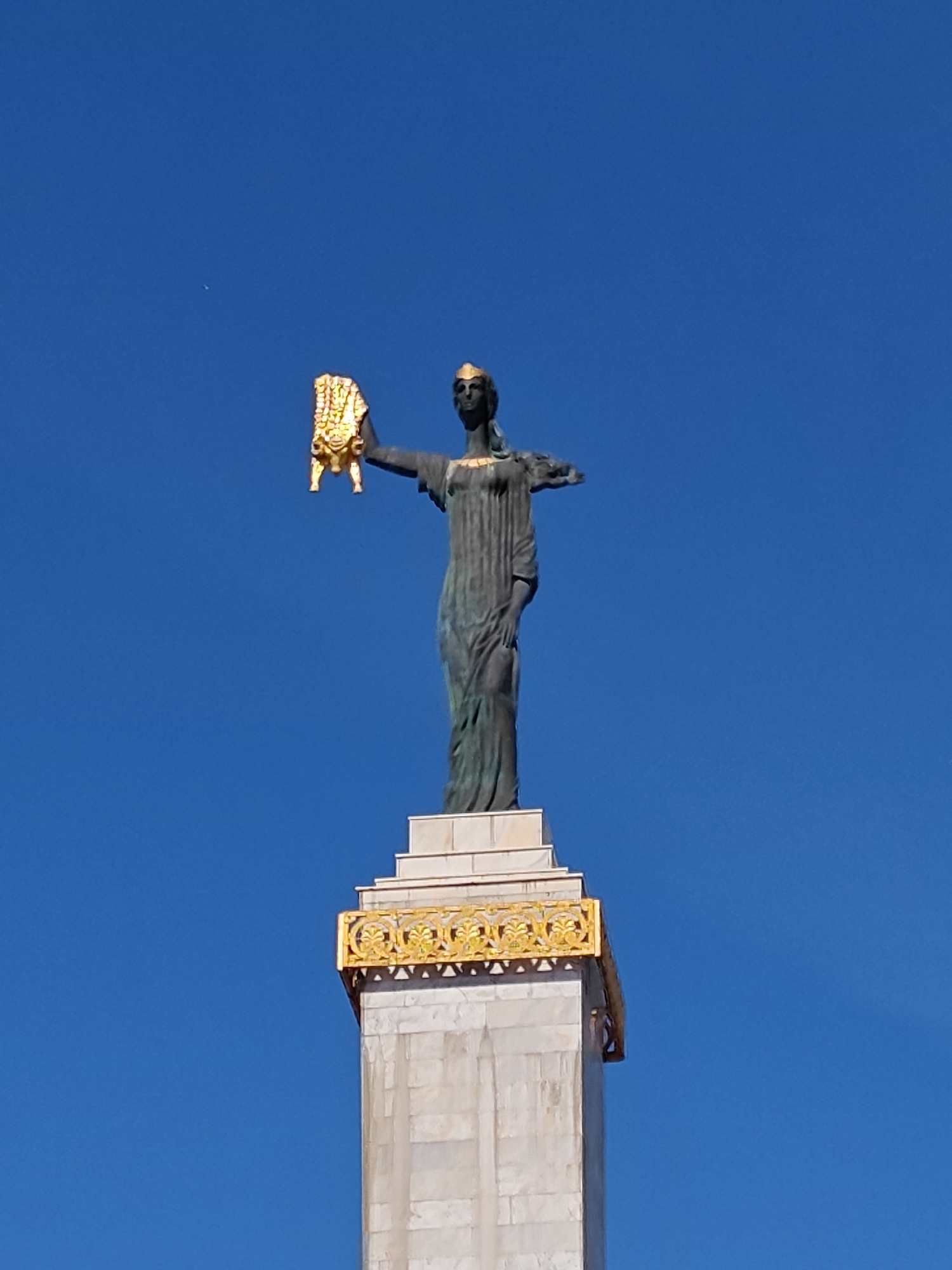 A bronze statue of Medea is atop a tall column with golden trim, set against a clear blue sky. The statue holds a golden fleece aloft in its right hand. The statue is located in the city of Batumi, Georgia.