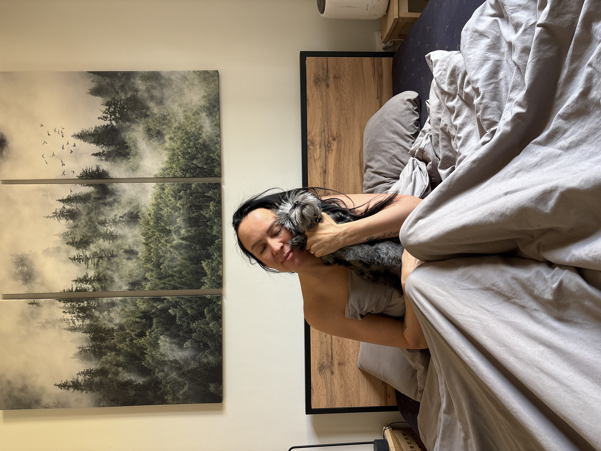 A woman is lying in bed holding a small dog. She is facing the camera with her eyes closed. The dog is gray and white and is nuzzling into her neck. The bed is covered in gray sheets and a gray pillow is visible. Behind the bed is a wooden headboard with a black frame. On the wall to the left of the bed is a triptych of a forest scene with fog and birds flying in the sky. A small wooden shelf with a white object on it is to the right of the headboard. The location is indoors and appears to be a bedroom. There is no indication of the city of Chaikovsky, Russia in the image.
