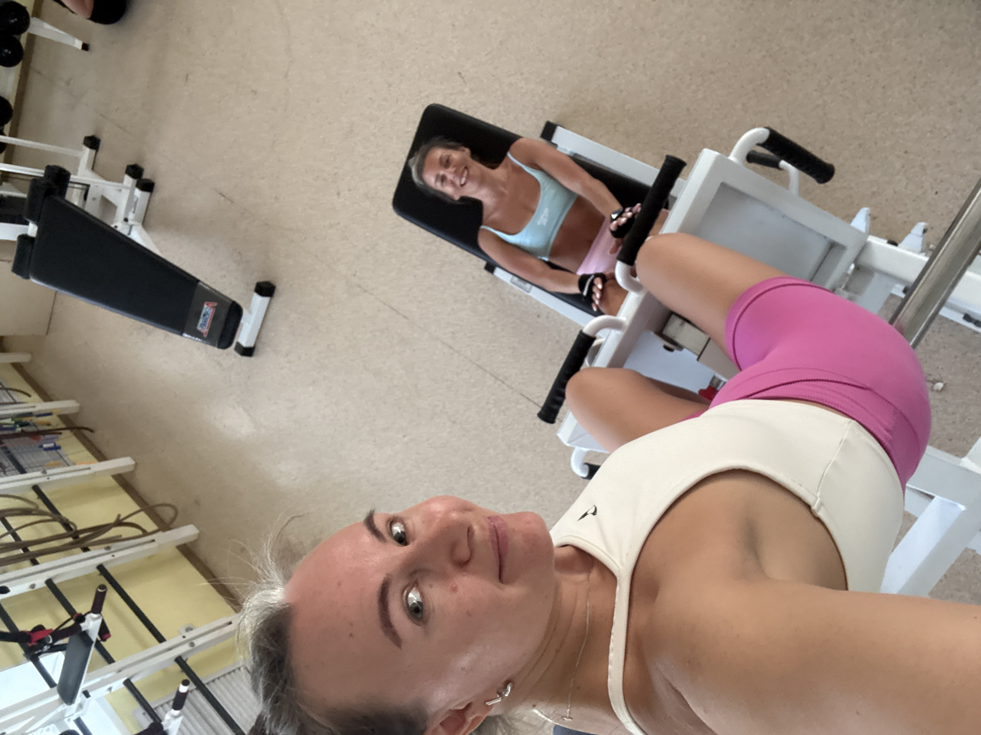 A selfie photograph taken indoors depicts two Caucasian women in workout attire. The foreground subject, closer to the camera, is wearing a white sports bra and bright pink shorts. Her hair is light-colored and pulled back, and she is wearing earrings and a necklace. She appears to be smiling slightly.
In the background, another woman is lying on a weight machine, wearing a light blue sports bra and pink shorts. She is smiling and looks towards the camera. The room seems to be a gym, with various exercise machines and equipment visible in the background.
Based on the visual information, the location is most likely a fitness center or gym in Chaikovsky, Russia. No interaction is taking place between the subjects.