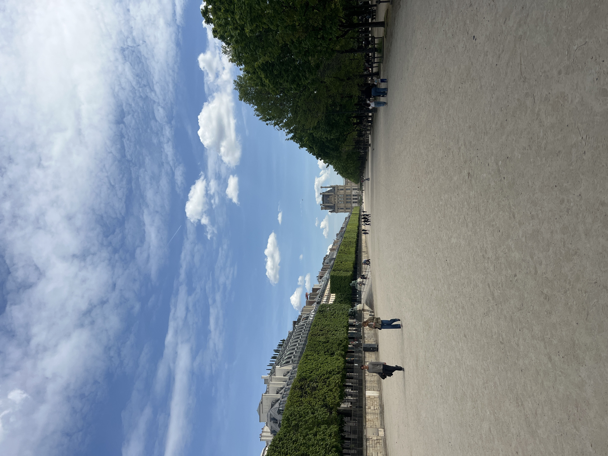 This outdoor image, taken in Paris, France, captures a wide, long perspective of what appears to be the Tuileries Garden, looking towards the Louvre Museum. The scene is dominated by a broad, light-colored pathway, likely made of dirt or gravel, stretching into the distance.
On the left side of the path, a dense line of tall, leafy green trees forms a grand avenue, with several park benches visible beneath their shade. On the right side, a series of elegant buildings with distinctive rooftops and green shrubbery or hedges below them frame the pathway. In the far distance, centered, stands a large, ornate architectural complex, strongly resembling the Louvre Museum.
The sky is a vibrant blue, adorned with scattered white clouds, ranging from fluffy cumulus to thinner, wispy cirrus formations. A faint airplane contrail is discernible in the upper sky. The weather appears sunny and pleasant, suggesting daytime, likely late morning or afternoon.
Several people are casually present in the scene. In the midground, on the right side of the path, two individuals are walking away from the viewer; one is dressed in a light-colored jacket and blue jeans, and the other in darker clothing. Other smaller figures can be seen walking or congregating further down the path and near the tree-lined avenue. The overall atmosphere is one of peaceful leisure in a grand public space. There is no visible text in the image.