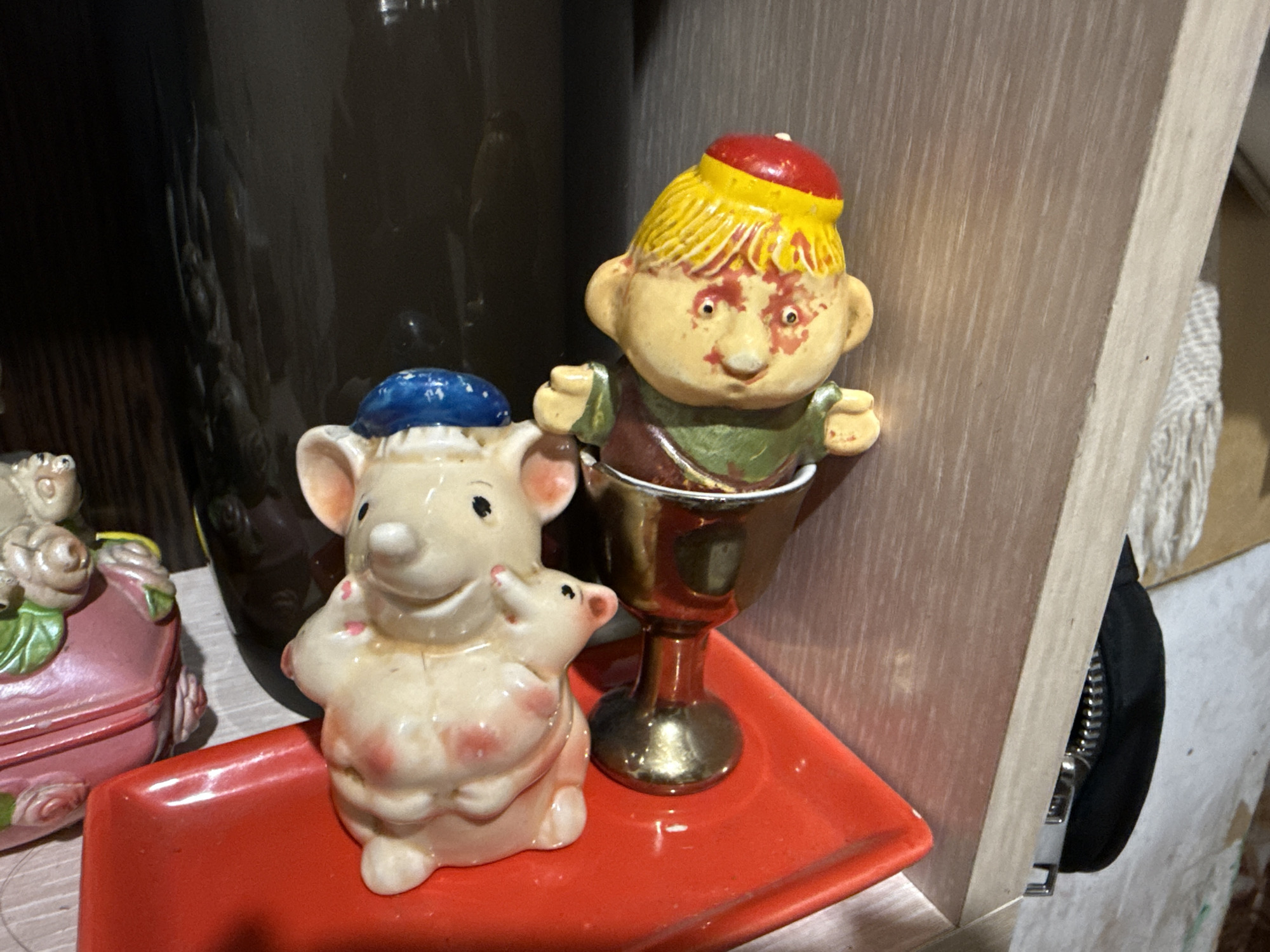 The image depicts a still life arrangement on a wooden surface, with a dark, cylindrical object in the background and a light-colored wooden board forming a corner on the right. In the foreground, a red, rectangular tray holds two ceramic figurines. On the left, a light-colored mouse figurine wearing a blue hat is shown holding a smaller mouse figurine. On the right, a ceramic figurine of a boy with yellow hair and a red hat is depicted inside a golden chalice-like object. The boy wears a green and brown tunic. The overall scene suggests an indoor setting, possibly a shelf or tabletop. The context of Nyurba, Russia, is not visually ascertainable from the provided image.