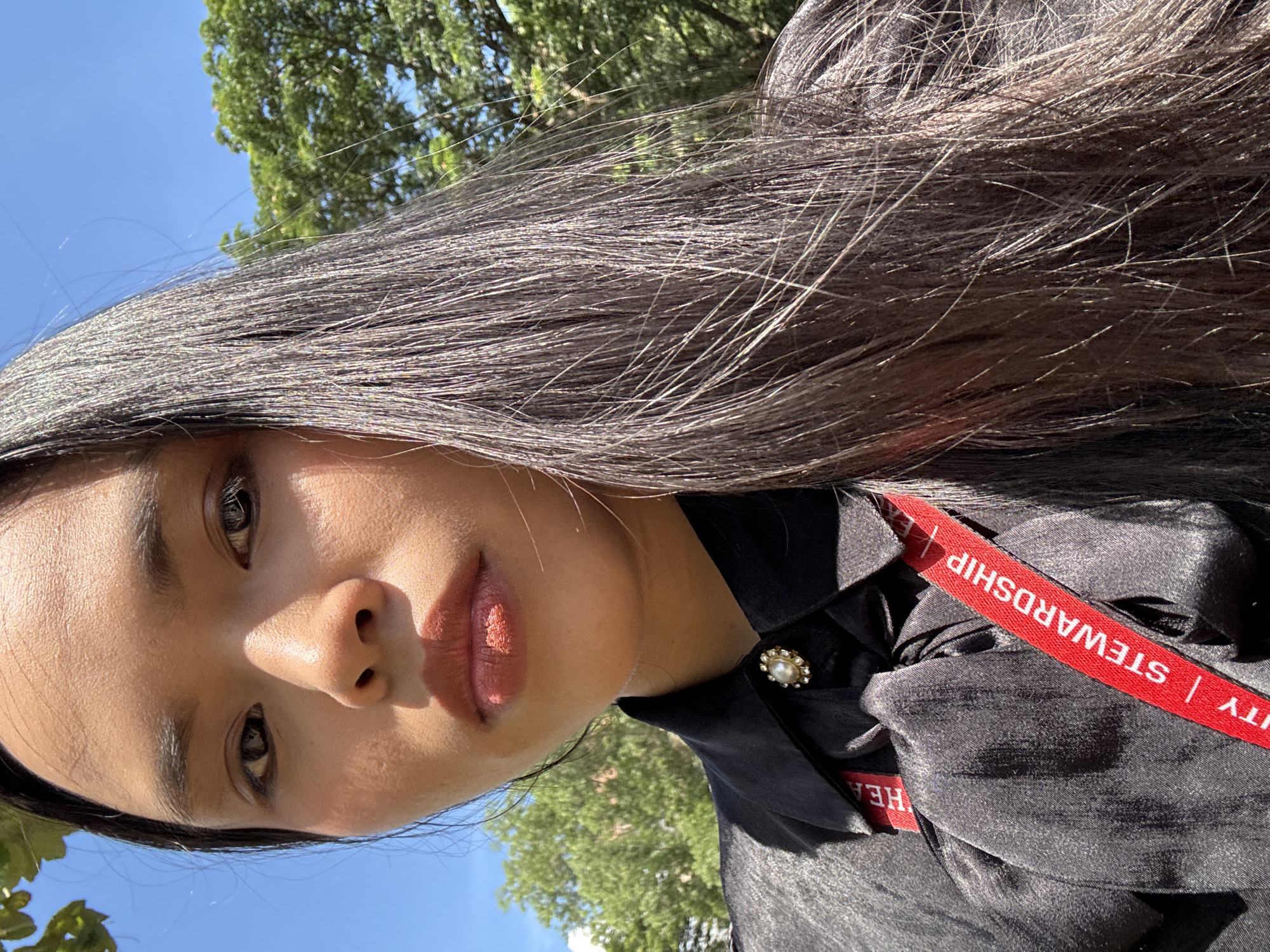 A medium, low-angle, eye-level outdoor photograph shows a light-skinned Asian female in Davao City, Philippines. She wears a black satin blouse fastened at the collar with a pearl and gold pin. A red lanyard with "STEWARDSHIP" printed in white is visible. The sky is blue, and green trees are in the background.