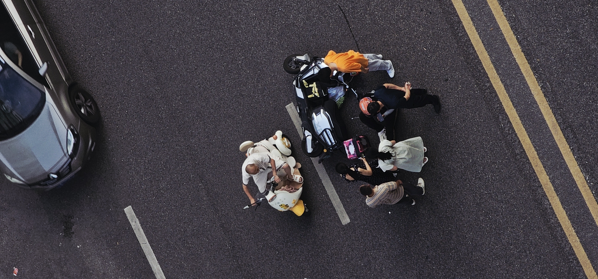 The image, taken from an elevated perspective, depicts a street scene in what appears to be Jiulongpo District, China. A gray car is positioned on the left side of the frame. Two motorcycles lie on the asphalt, surrounded by several people. One motorcycle is black and white, with its luggage carrier possibly containing delivery items. A group of people are gathered around it, some wearing an orange shirt and light-colored pants, a black shirt and black pants, a light green dress, and a striped shirt with dark pants. Nearer to the left, a second, smaller white and yellow motorcycle is ridden by a man wearing white clothes. Road markings, including a solid white line and double yellow lines, are visible on the asphalt surface.