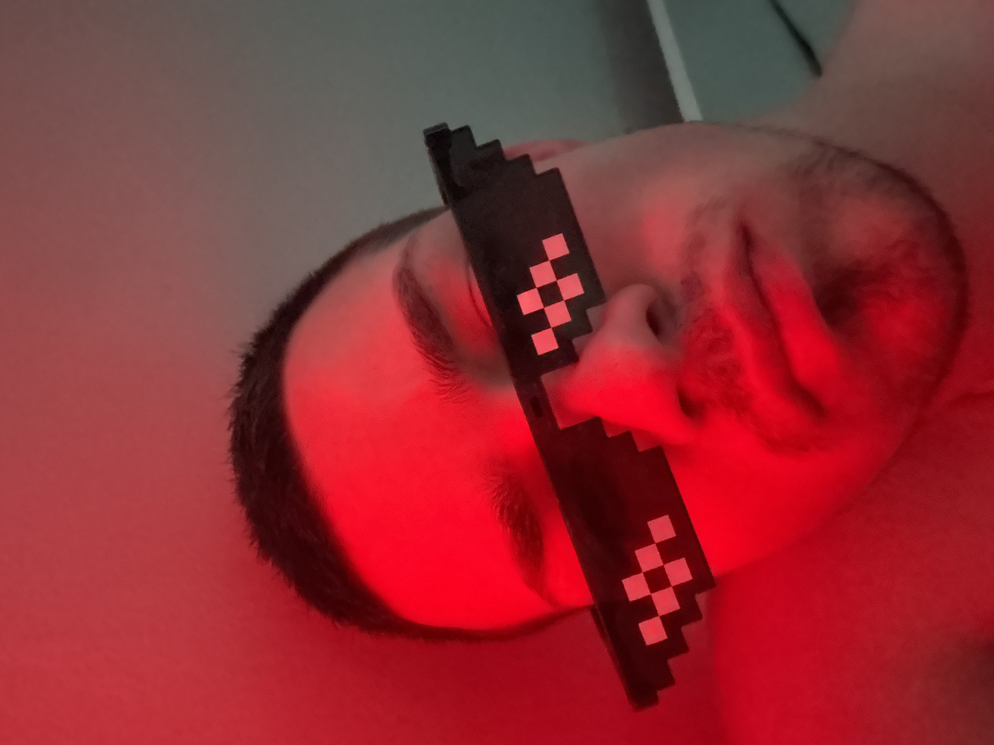 A close-up, low-angle selfie captures a person wearing pixelated sunglasses, lit by a strong red light. The person's face, from the forehead to the chin, is visible, with their eyes obscured by the sunglasses. The background is a plain, textured surface, possibly a wall, illuminated with a gradient of red light. The overall impression is one of a posed, informal shot, likely taken indoors. The specific location in the city of Varna, Bulgaria, cannot be determined from the visual information provided.