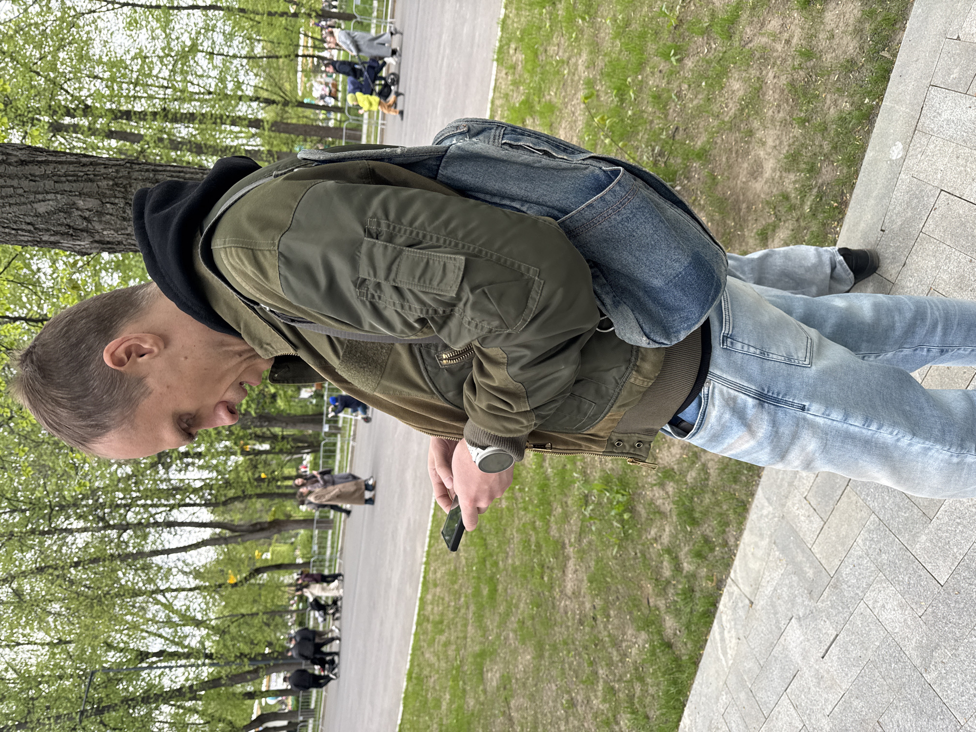 A person in a green jacket and jeans is standing in a park, holding a cell phone. The park appears to be in Moscow, Russia. The background shows trees, grass, and a paved walkway with people. The person is wearing a watch. The weather seems mild.