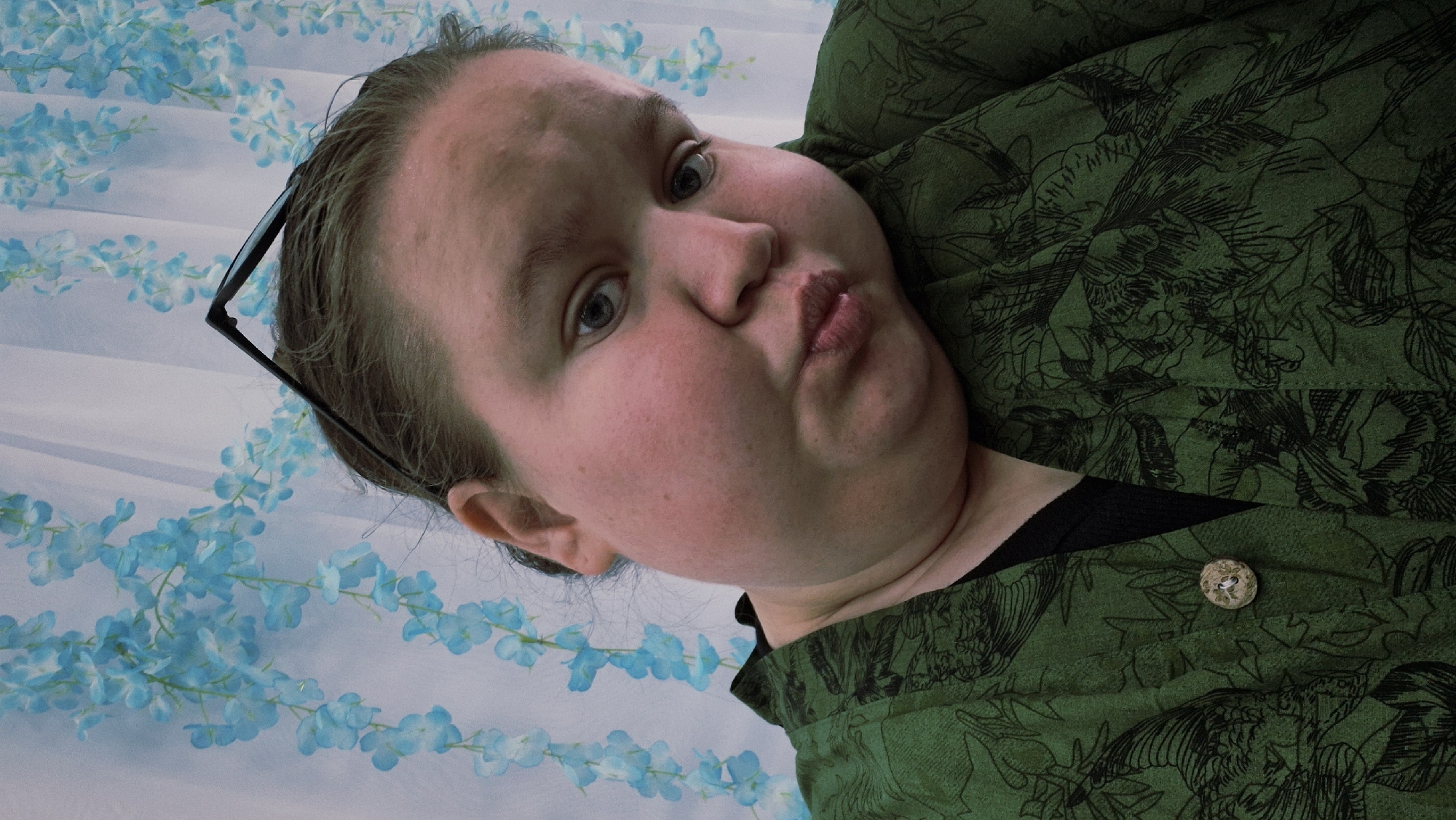 An eye-level shot captures a person, appearing to be an adult female, with fair skin and light brown hair pulled back, with black-framed glasses resting on her head. Her face fills a significant portion of the frame, angled as if looking towards the camera. She has light-colored eyes (possibly blue or grey) and her lips are pursed or puckered in a playful expression. Her cheeks are full and flushed.
She is wearing a dark green garment, possibly a jacket or top, with a repeating dark black floral or leafy pattern. A small, light-colored button with a textured, possibly organic, appearance is visible on the green fabric near the bottom right. A black undergarment is visible beneath the green top near her neck.
The background consists of a light, possibly white or very light blue, fabric or wall adorned with strands of light blue artificial flowers or vines. The entire image is rotated approximately 90 degrees counter-clockwise, making the person's head appear tilted upwards relative to the frame. The lighting is soft and even, suggesting an indoor setting.
There are no visible texts or specific location cues within the image that point to Chaikovsky, Russia, though that is the provided context. The activity is clearly posing for a photograph, likely a selfie, given the close-up and direct gaze. The person's expression conveys a sense of lightheartedness or amusement.