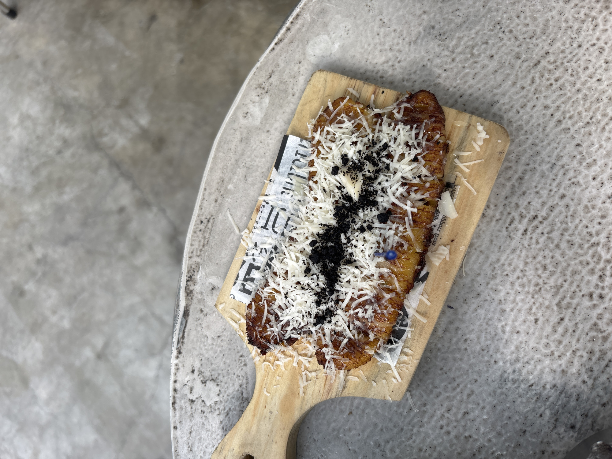 The image shows a top-down view of a wooden serving board holding a fried banana dessert, topped with shredded cheese and dark, granular crumbs (possibly chocolate or Oreo crumbs). The dessert is placed on a piece of newspaper or printed paper beneath the food. The serving board rests on a grey, textured surface that appears to be a tabletop. The location is likely North Jakarta, Indonesia, based on contextual information not directly visible in the image.
