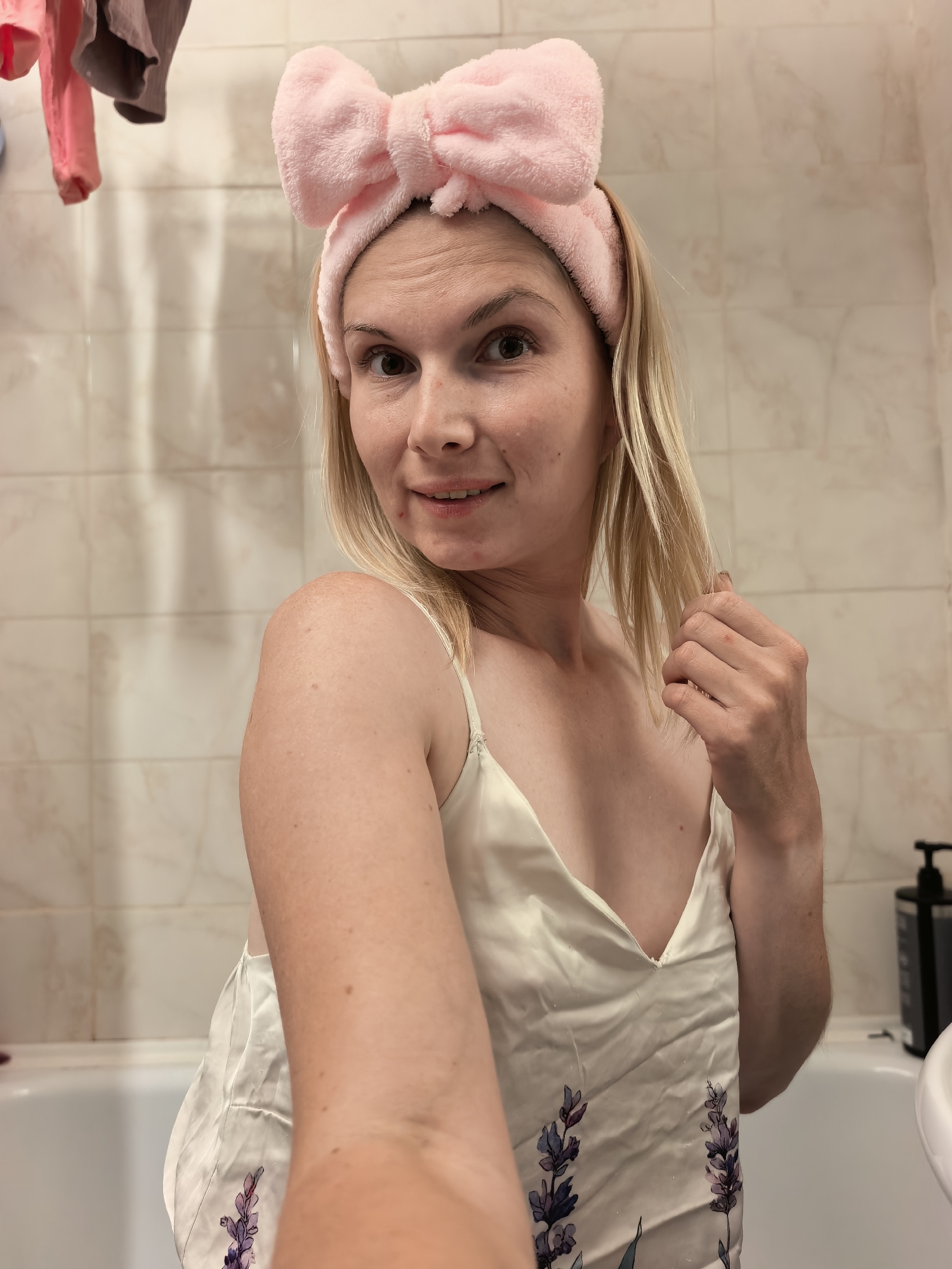 The image shows a blonde woman with a pink fluffy bow headband in a bathroom. She is wearing a white slip with a floral pattern. The bathroom has tiled walls and a bathtub. She is holding her hair with her right hand and smiling at the camera. The lighting suggests it is daytime, and the scene is indoors. There is a black pump bottle on the edge of the bathtub. There is no visible text in the image.
