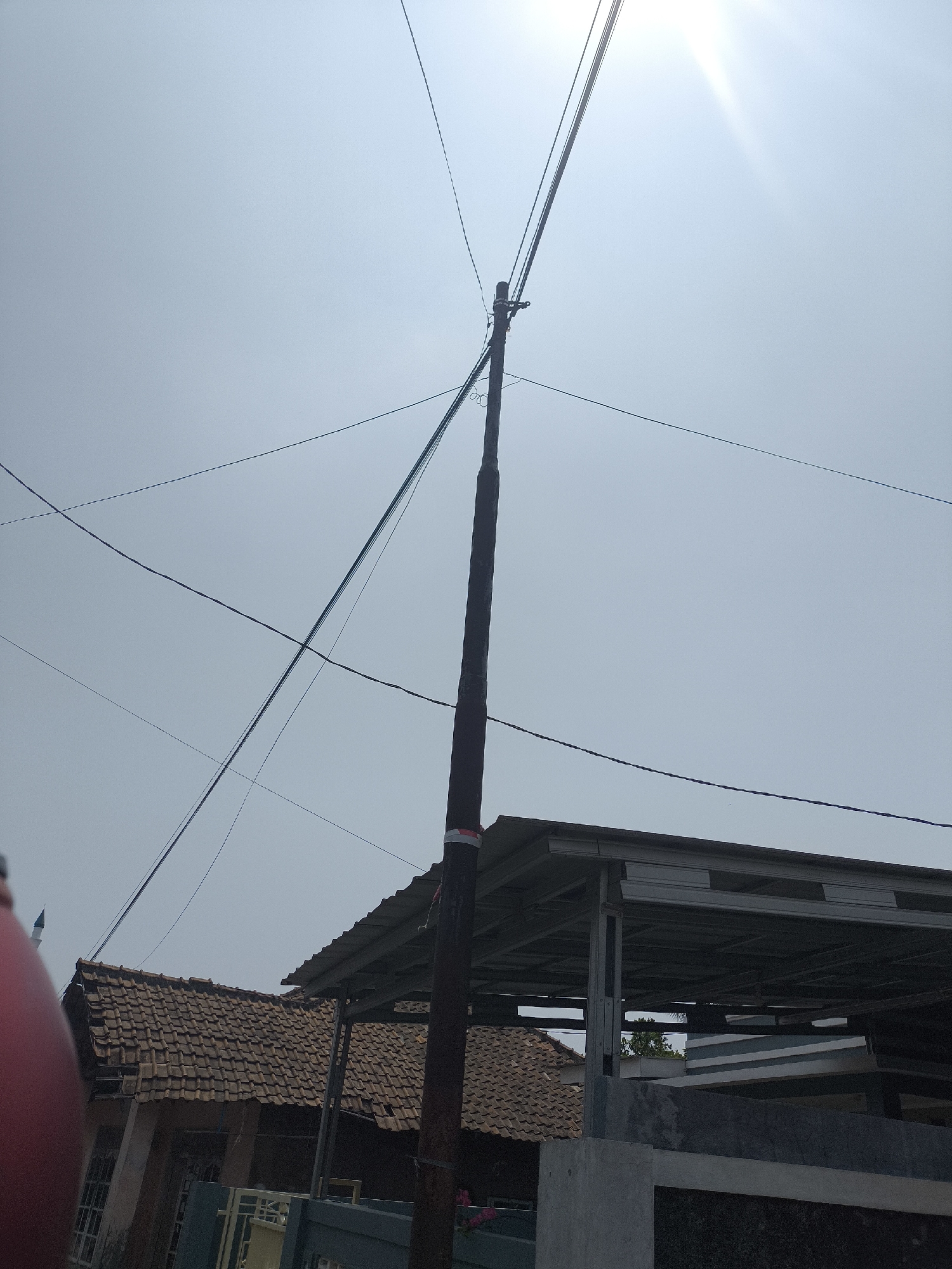 The image captures a view of overhead utility wires against a hazy sky, with a utility pole in the foreground. The pole, made of wood, is dark brown and has a red and white band wrapped around its upper section. Several electrical wires crisscross the sky, some taut and others with a slight sag. Below the wires, a portion of a building with a tiled roof is visible on the left, appearing to be a residential structure. To the right, a modern-looking building with a corrugated metal roof and a skeletal metal frame structure is present. The background consists of a light blue sky with a bright sunbeam emanating from the upper right corner, suggesting it is daytime. The overall scene suggests an outdoor environment in an urban or semi-urban setting, likely in Cilegon, Indonesia, given the context provided, potentially depicting the electrical infrastructure common in such areas.