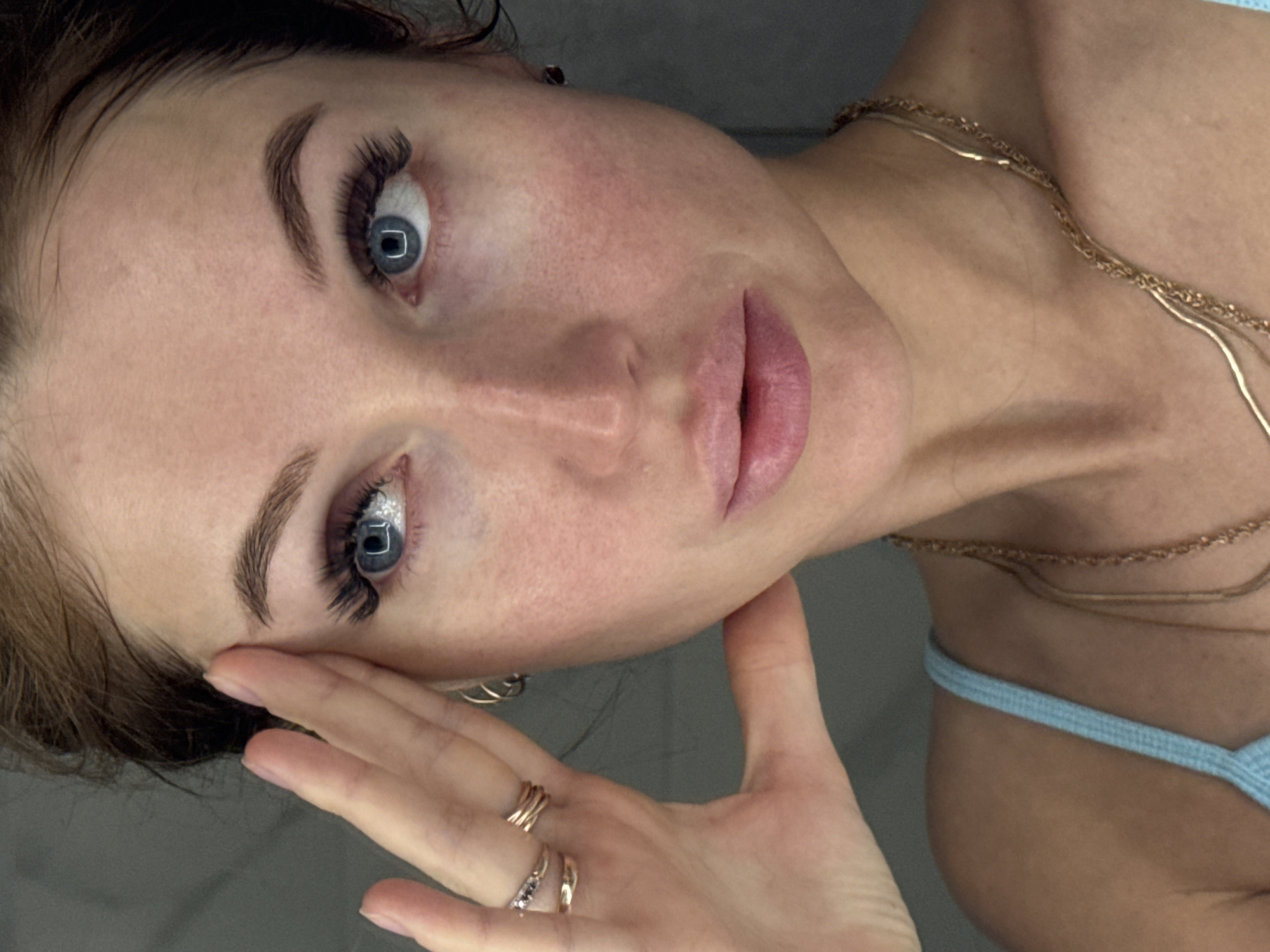 A close-up, rotated image shows a fair-skinned woman with blue eyes and dark eyelashes. She has well-defined eyebrows and pink lips. She is wearing gold necklaces and a light blue top, and her hand with gold rings is near her face. The background is a neutral gray. There is no indication of location in Chaikovsky, Russia.